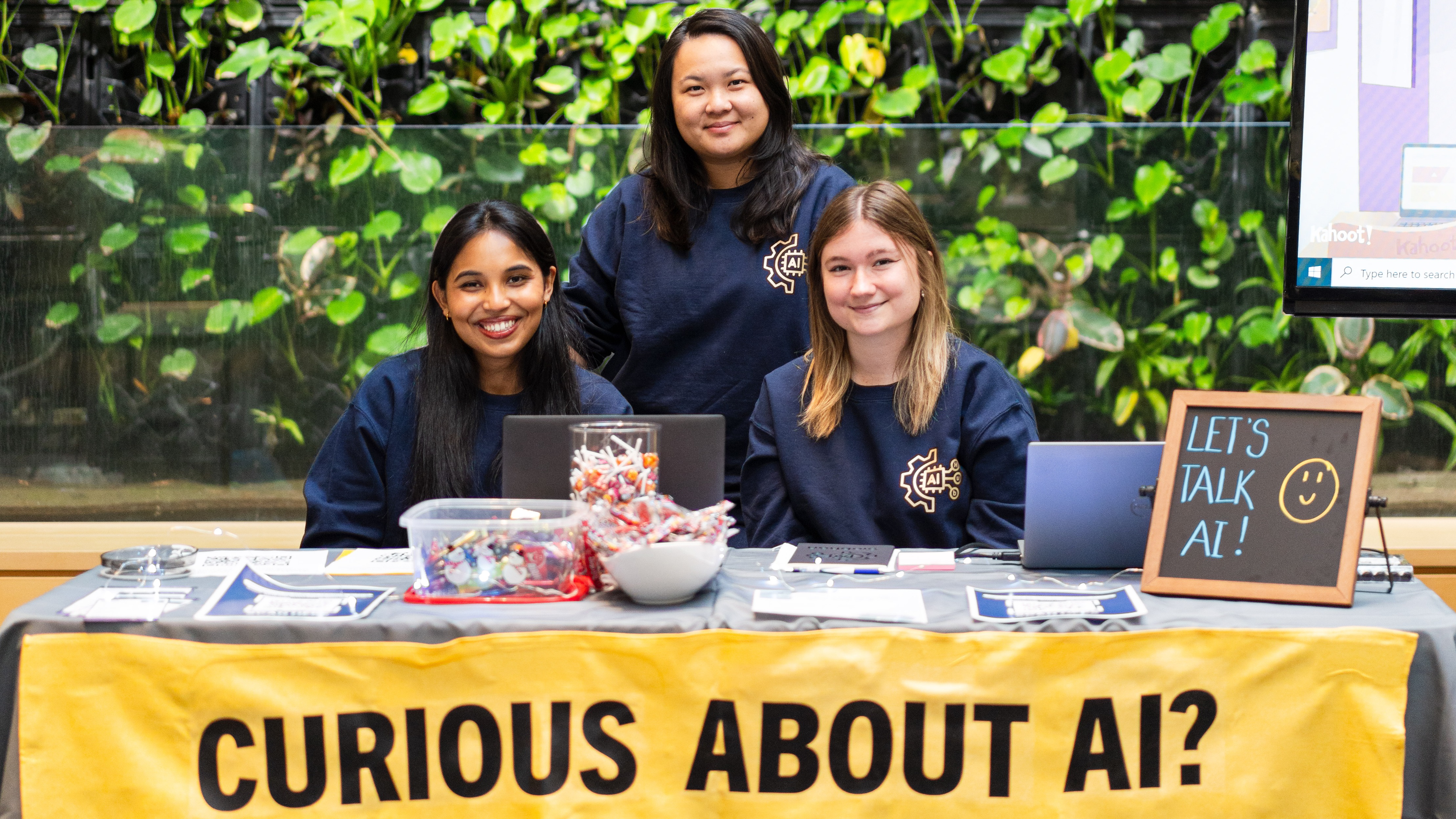 Three members of the AI Hub pose behind a table with a banner that reads CURIOUS ABOUT AI?