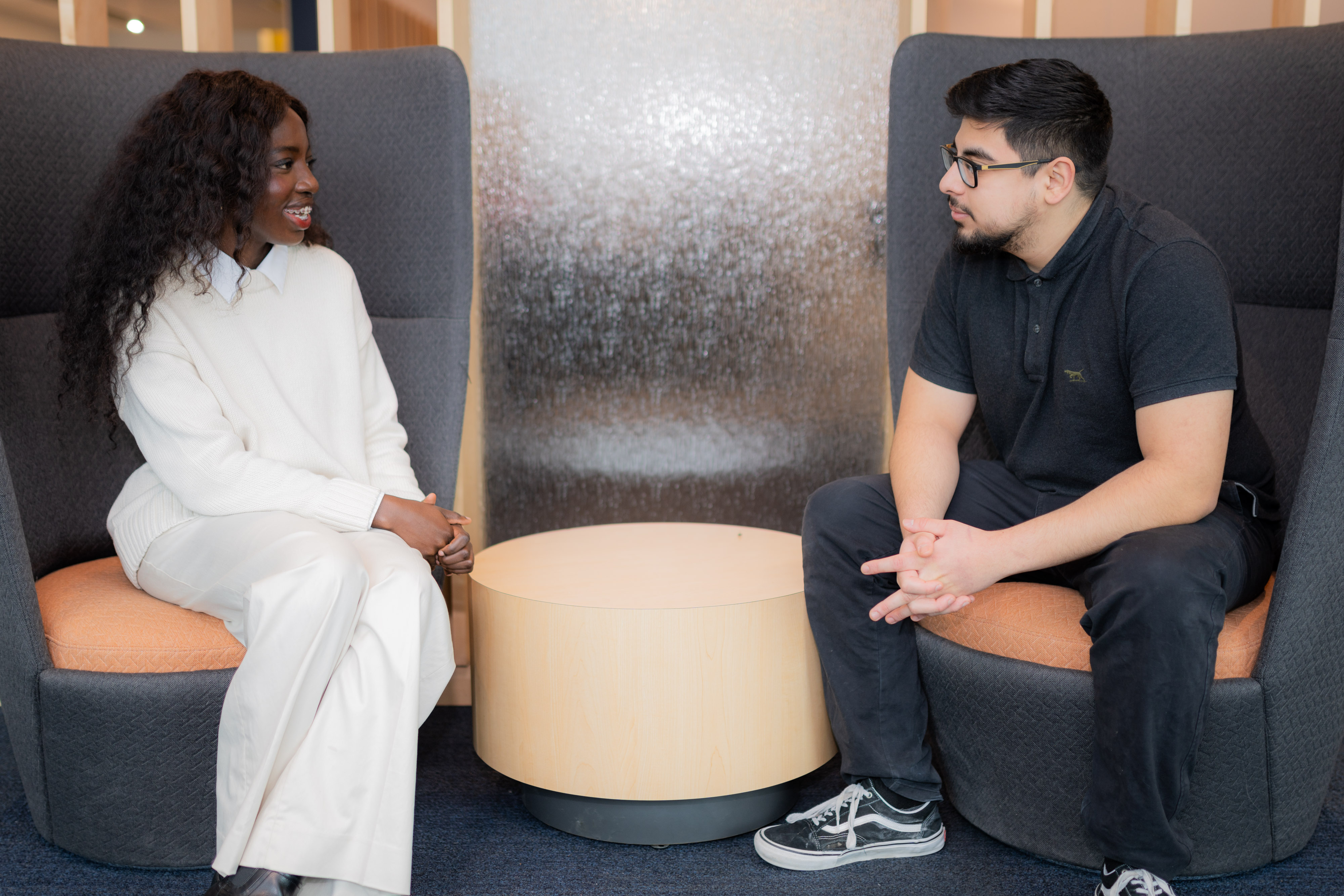 Two students sit on grey chairs having a discussion