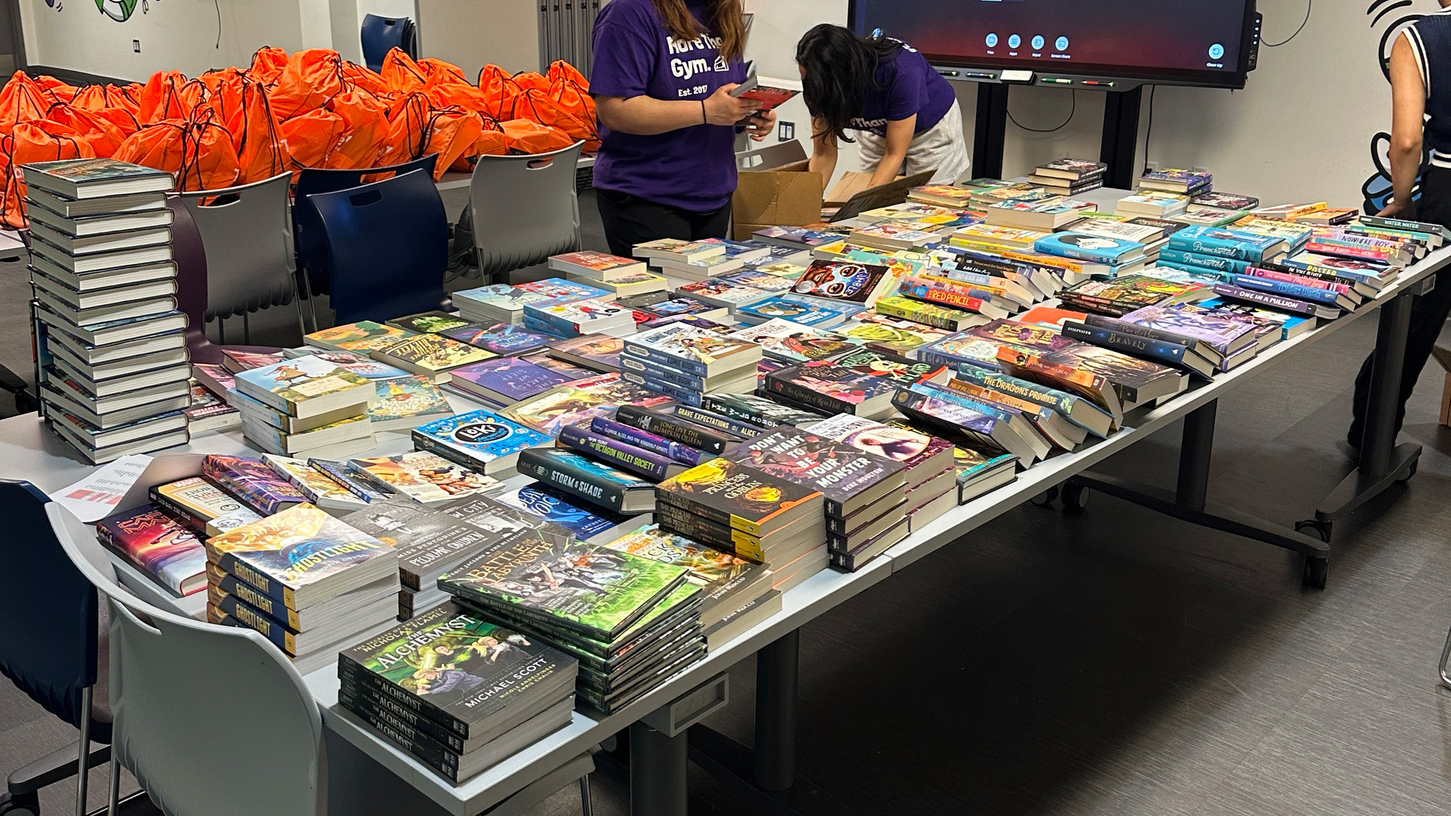 A long table filled with piles of books with two individuals organizing them by title