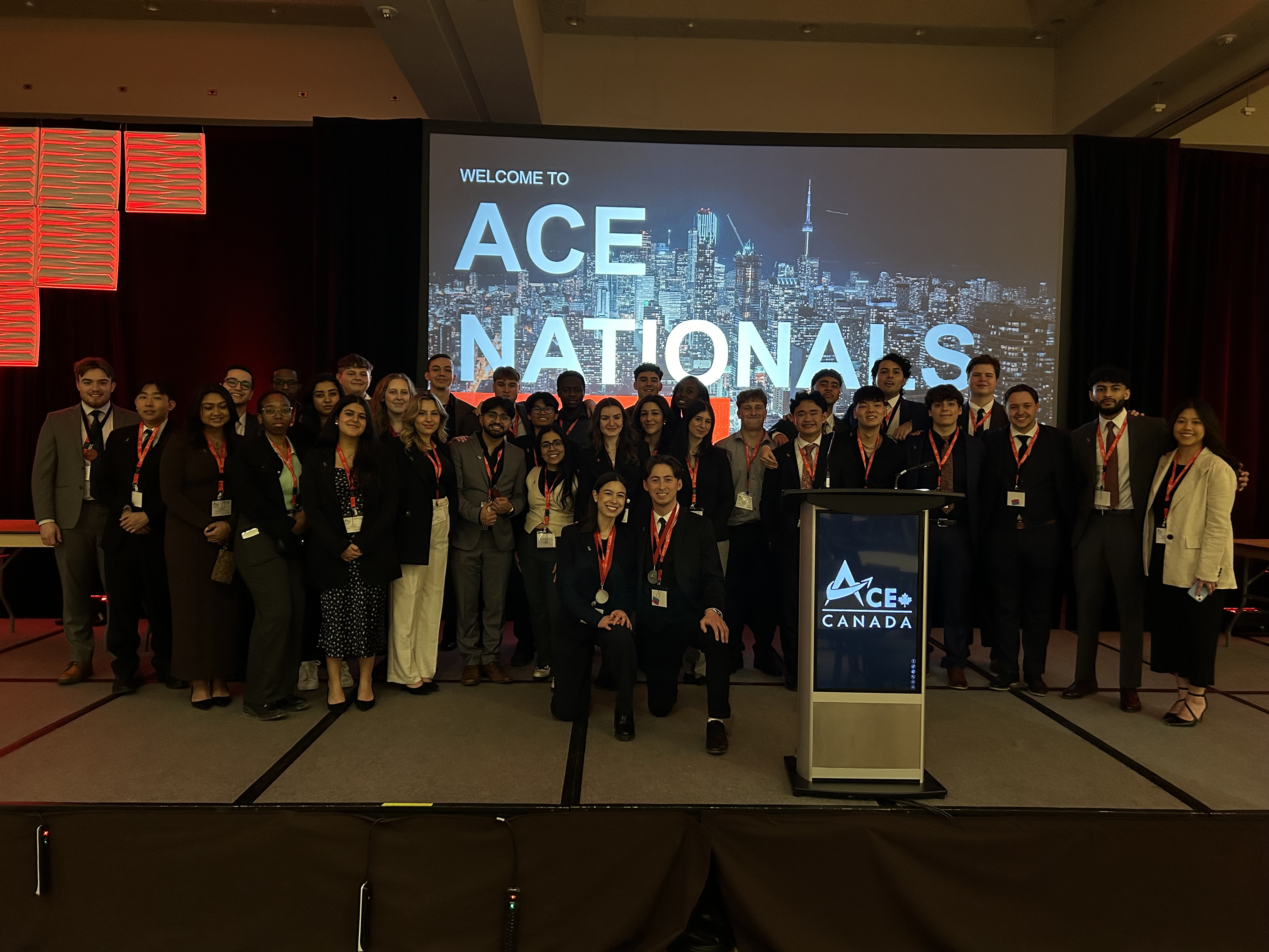 A large group of U of GH ACE students pose together in front of an ACE Nationals sign