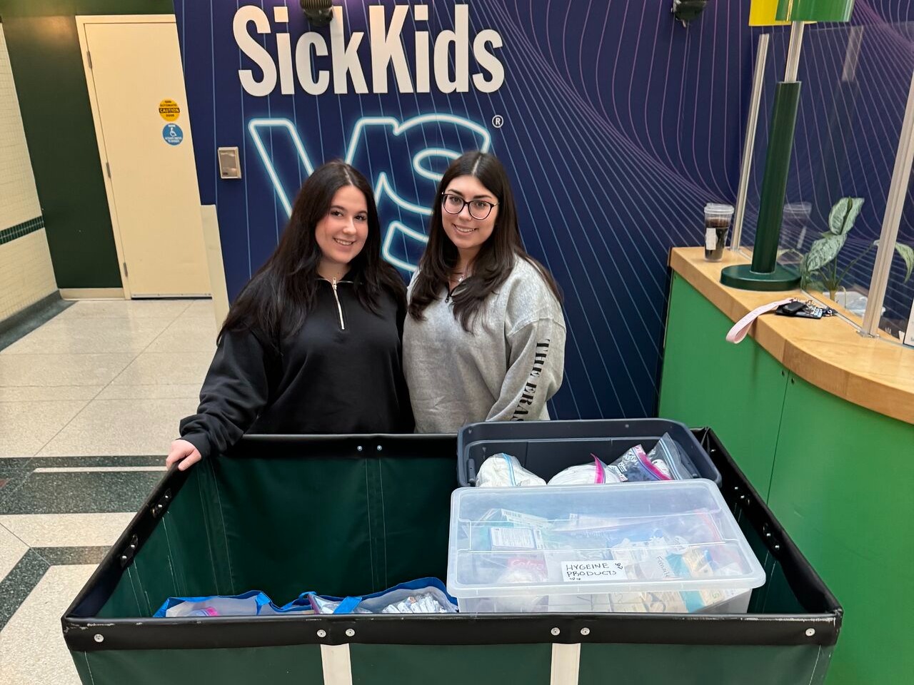 Two students pose in front of a SickKids sign with a bin of donations