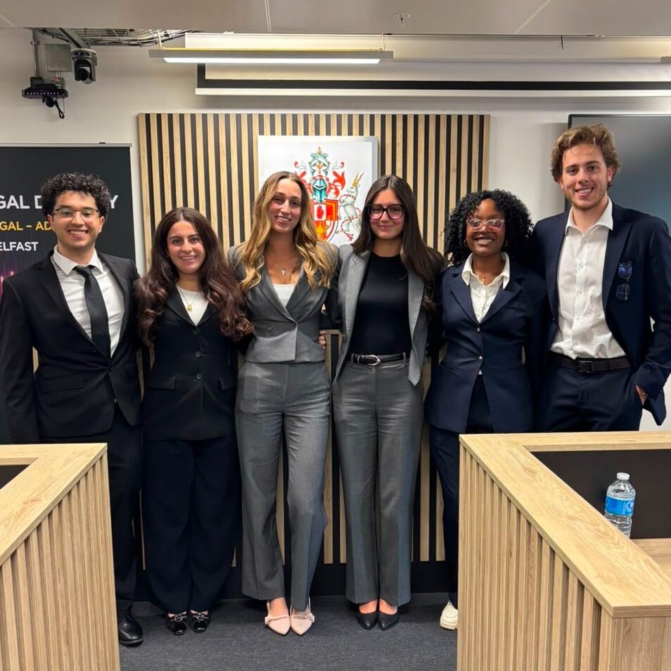 Six students stand at the front of a mock courtroom wearing suits