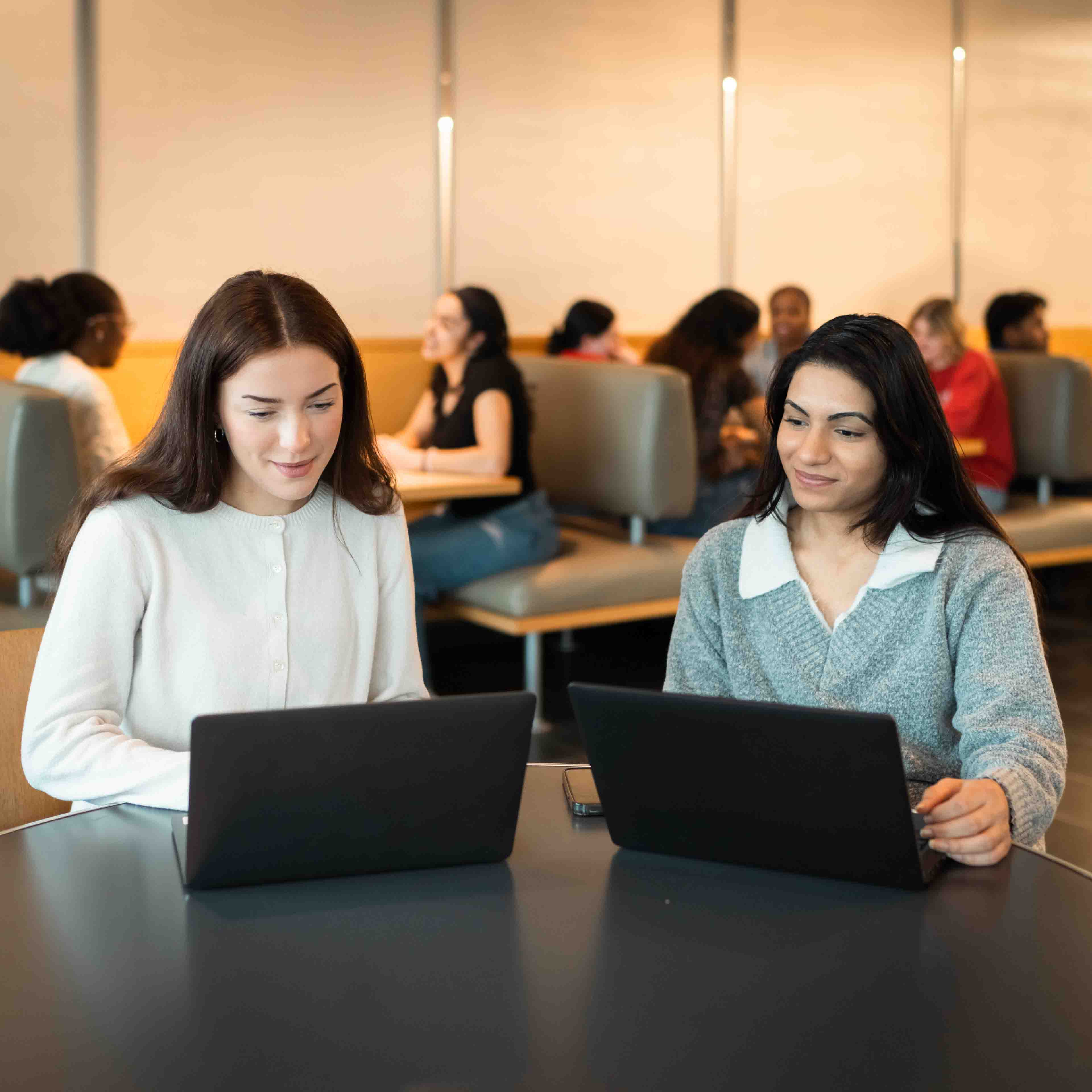 Two students sit around at a table with laptops