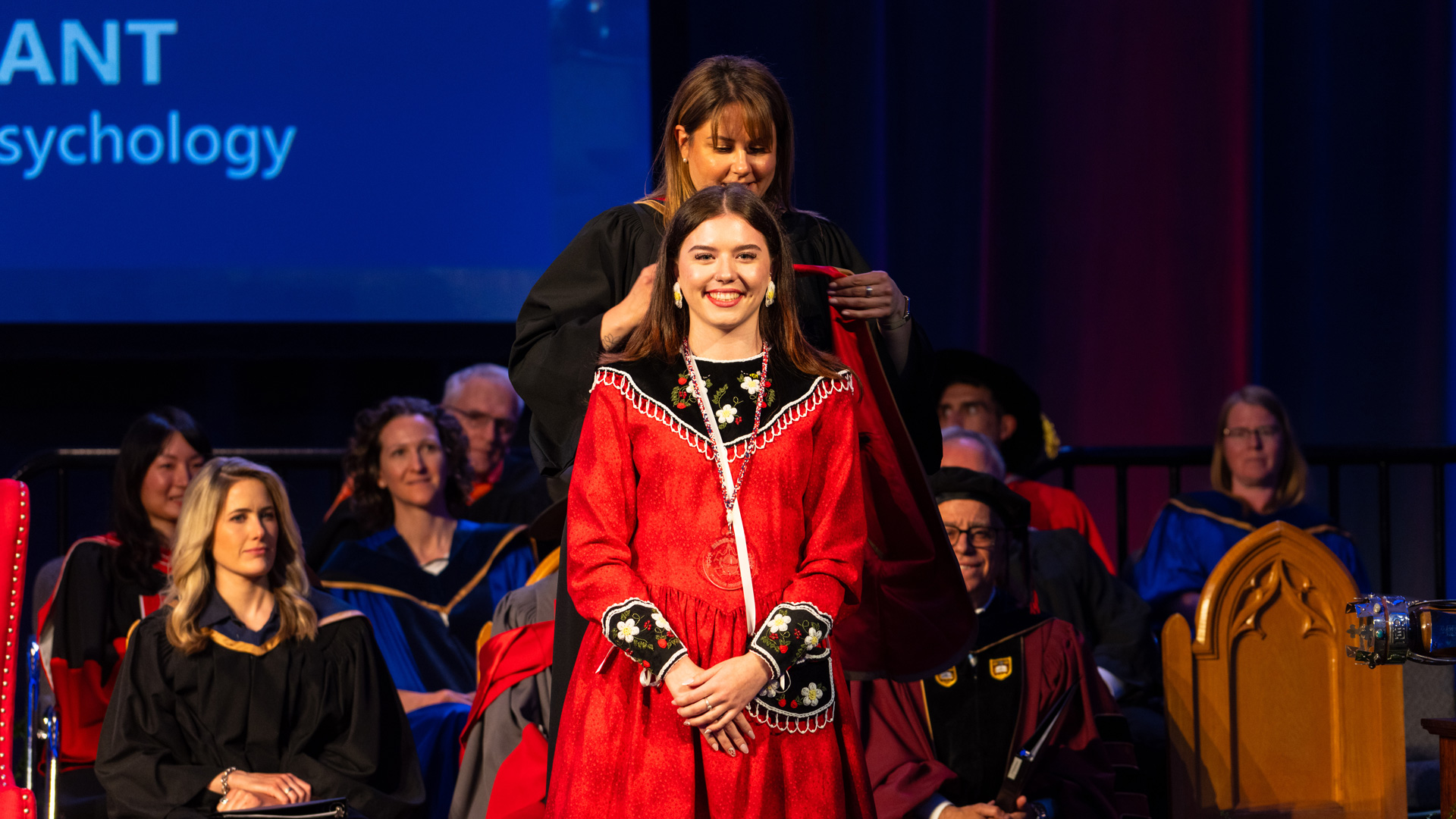 Kristen Brant in traditional Indigenous clothing smiling while waiting for someone to put the University of Guelph-Humber convocation hood over her. There are people in convocation regalia sitting behind, watching.