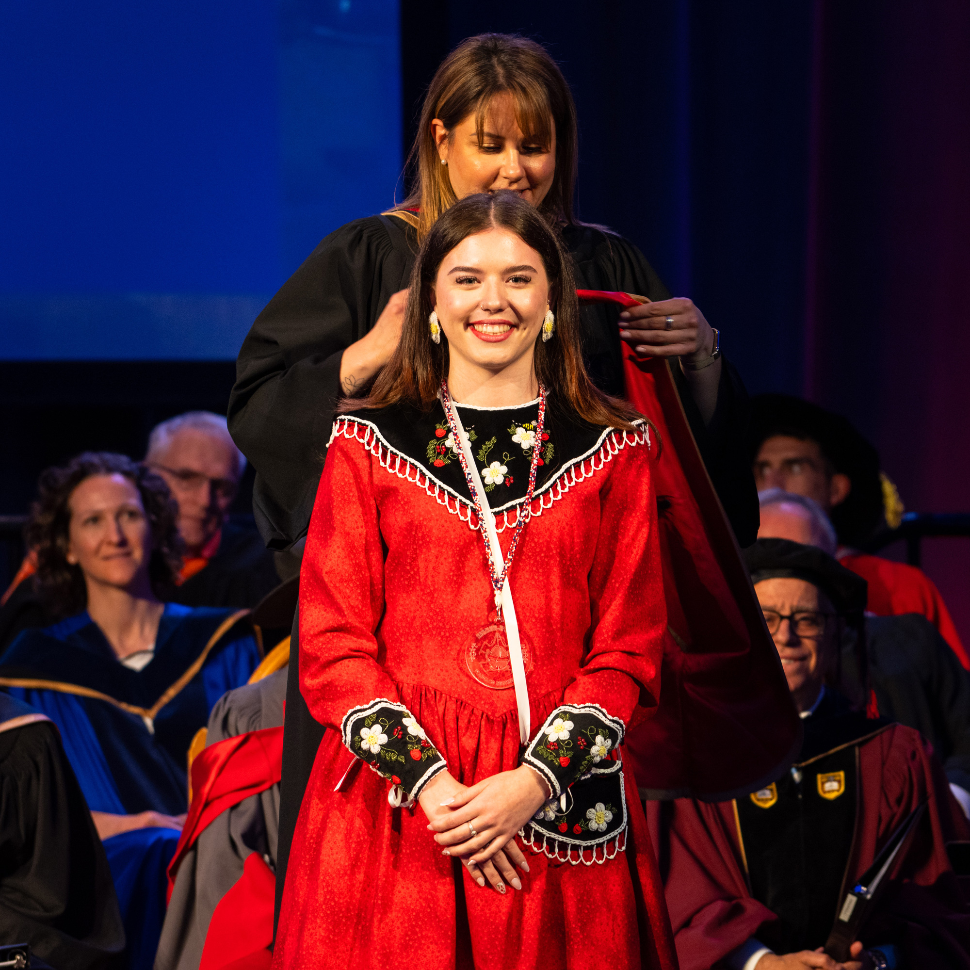 Kristen Brant in traditional Indigenous clothing smiling while waiting for someone to put the University of Guelph-Humber convocation hood over her. There are people in convocation regalia sitting behind, watching.