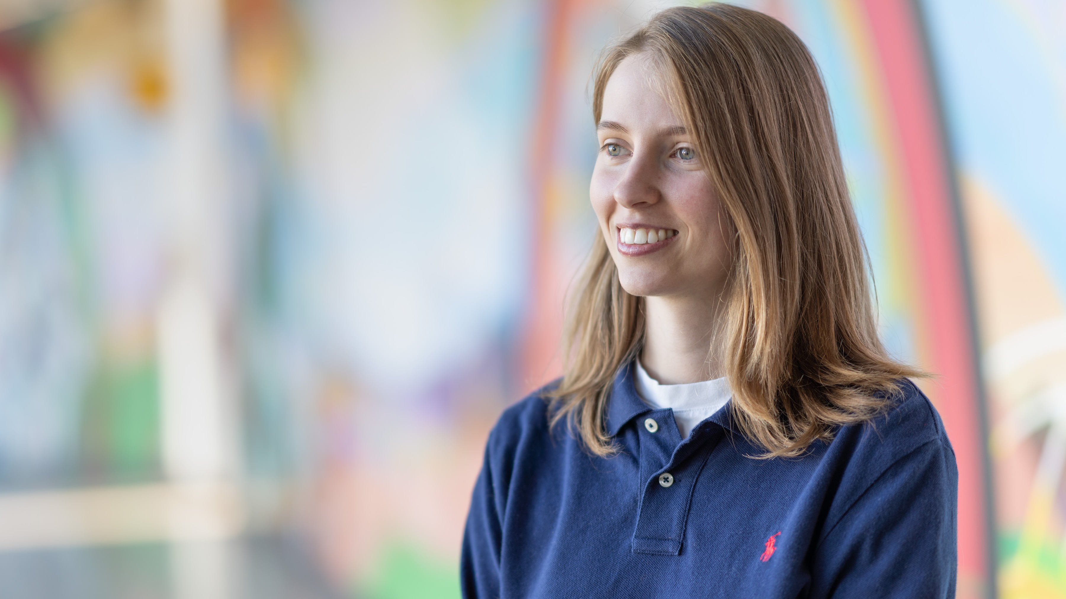 A woman, Kate Carveth, wearing a navy blue shirt poses in front of a colourful wall