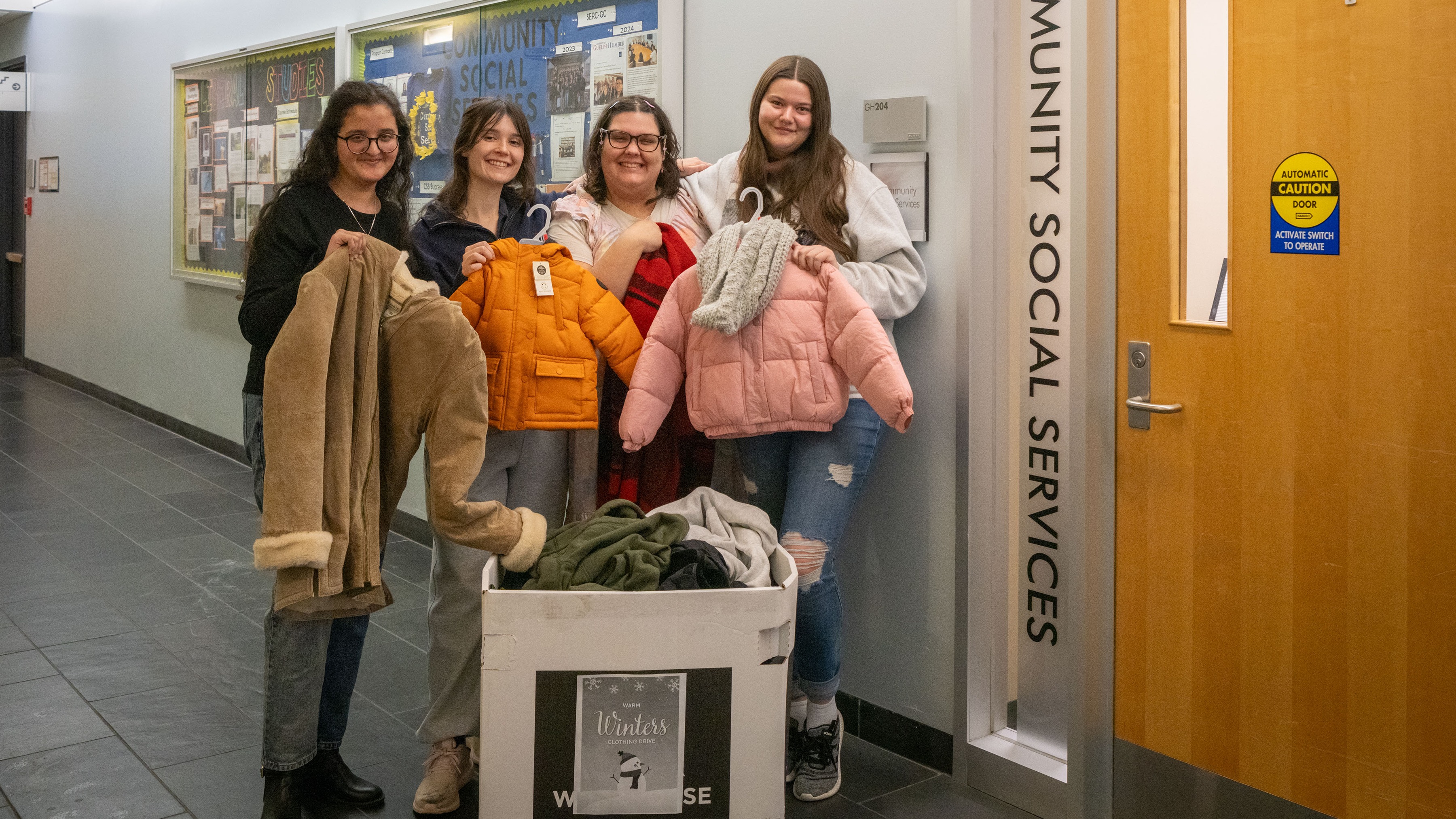 A group of four students holding jackets pose in front of a donation box by the Community Social Services office door