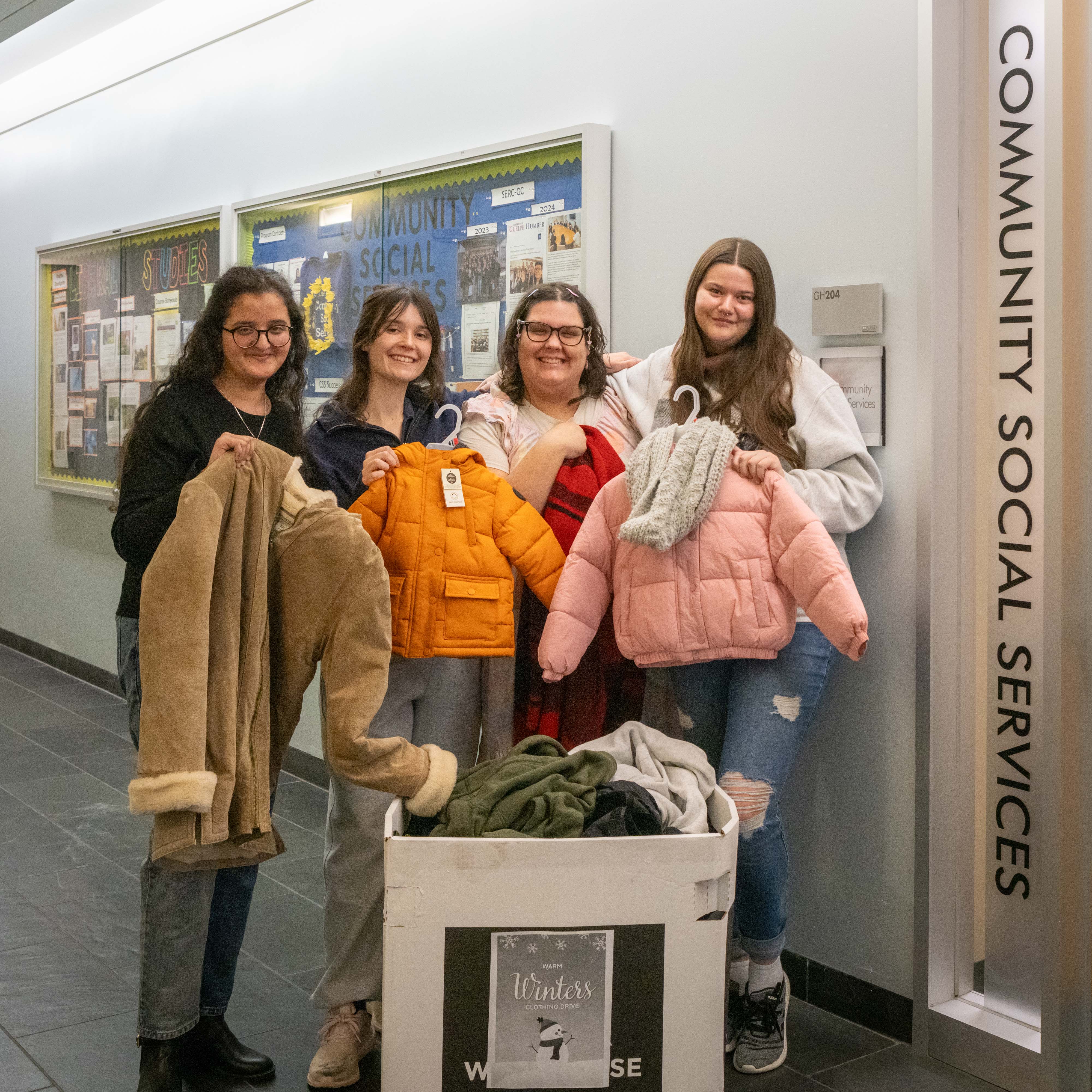 A group of four students holding jackets pose in front of a donation box by the Community Social Services office door