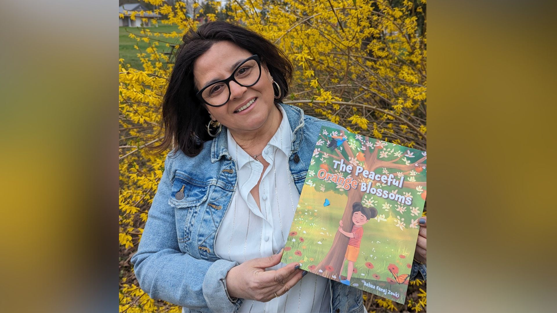 Bahaa poses with her book, The Peaceful Orange Blossoms, in front of a tree with yellow leaves