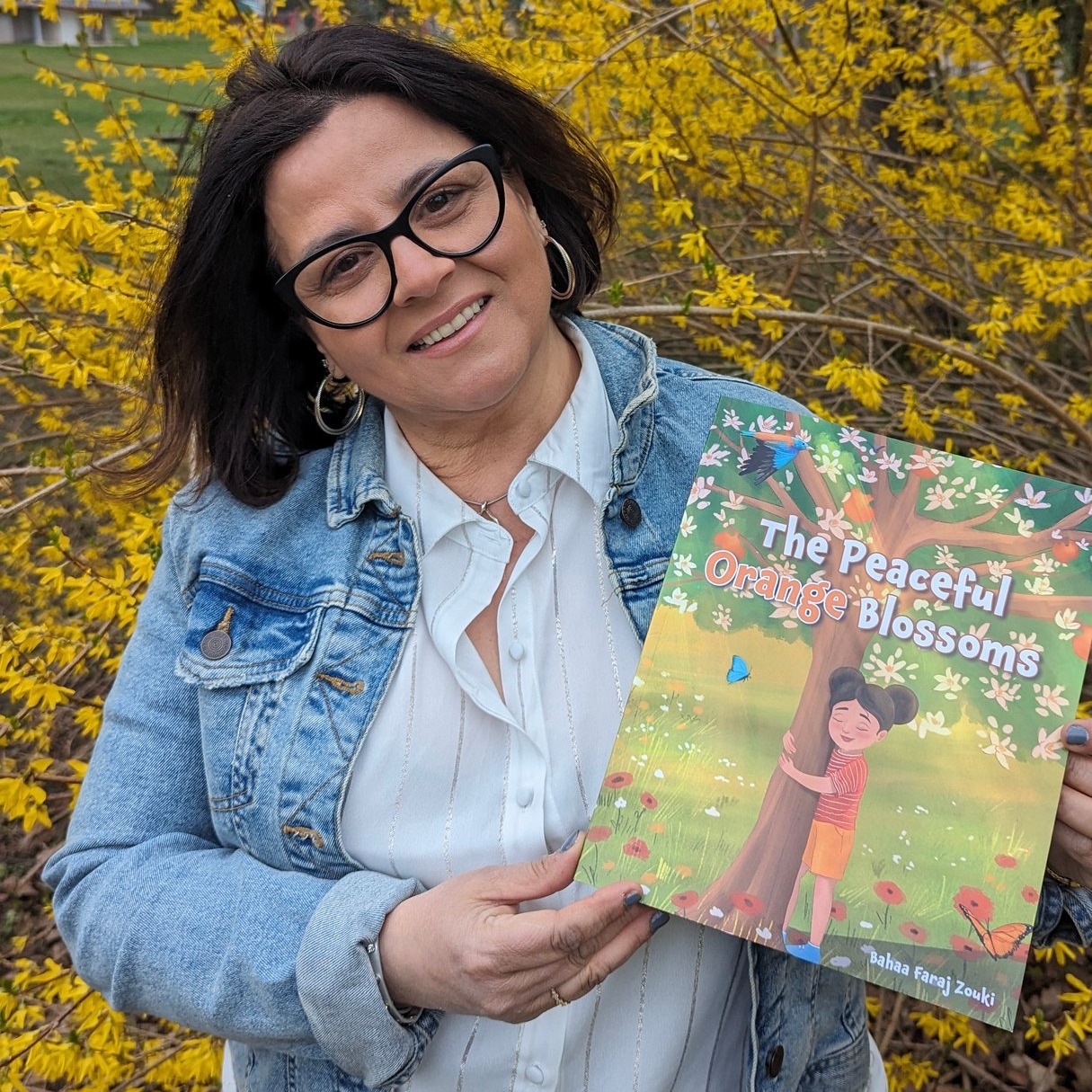 Bahaa poses with her book, The Peaceful Orange Blossoms, in front of a tree with yellow leaves