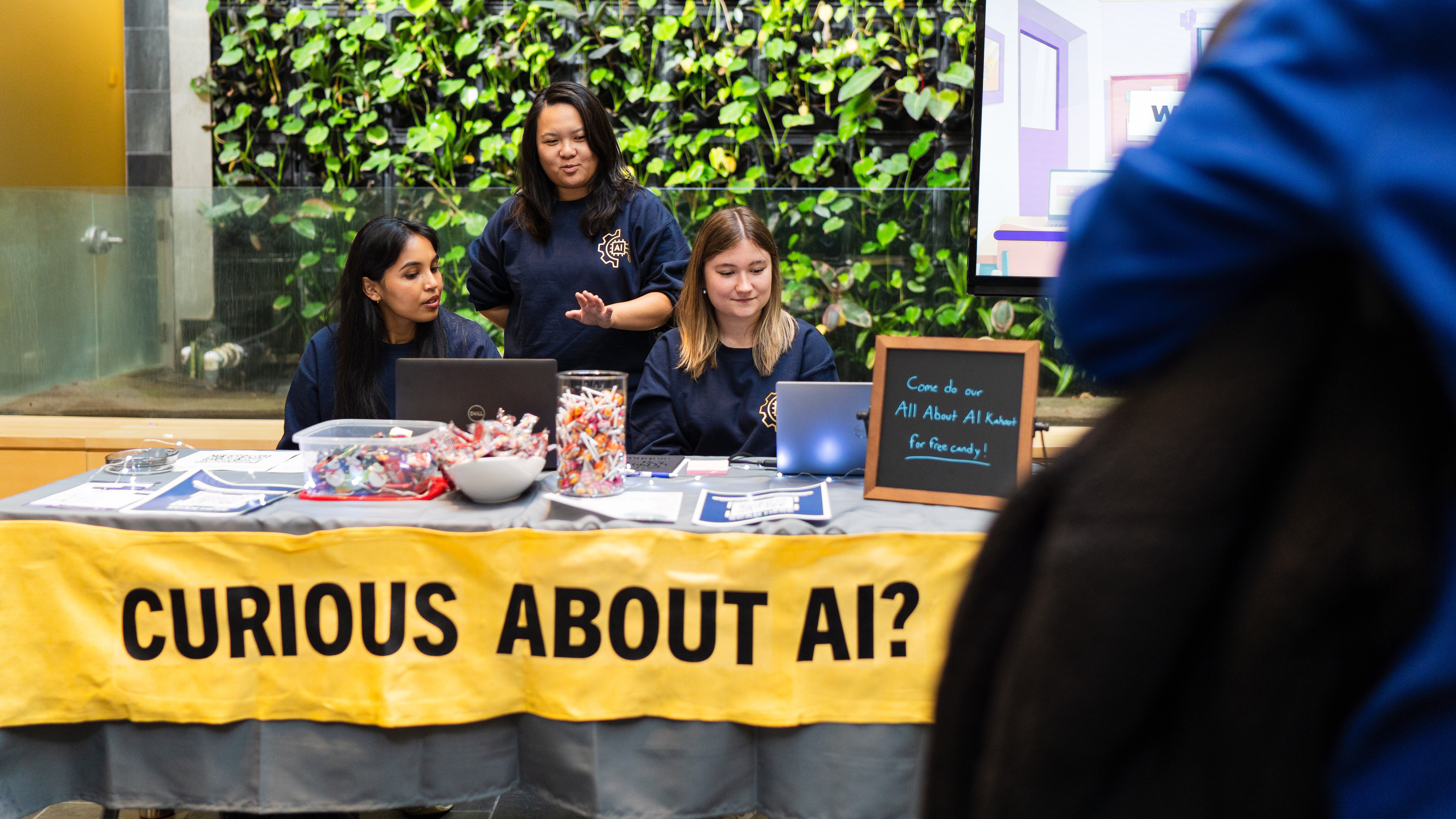 A group of three people forming the AI Hub sit by a table with a yellow sign that reads CURIOUS ABOUT AI?