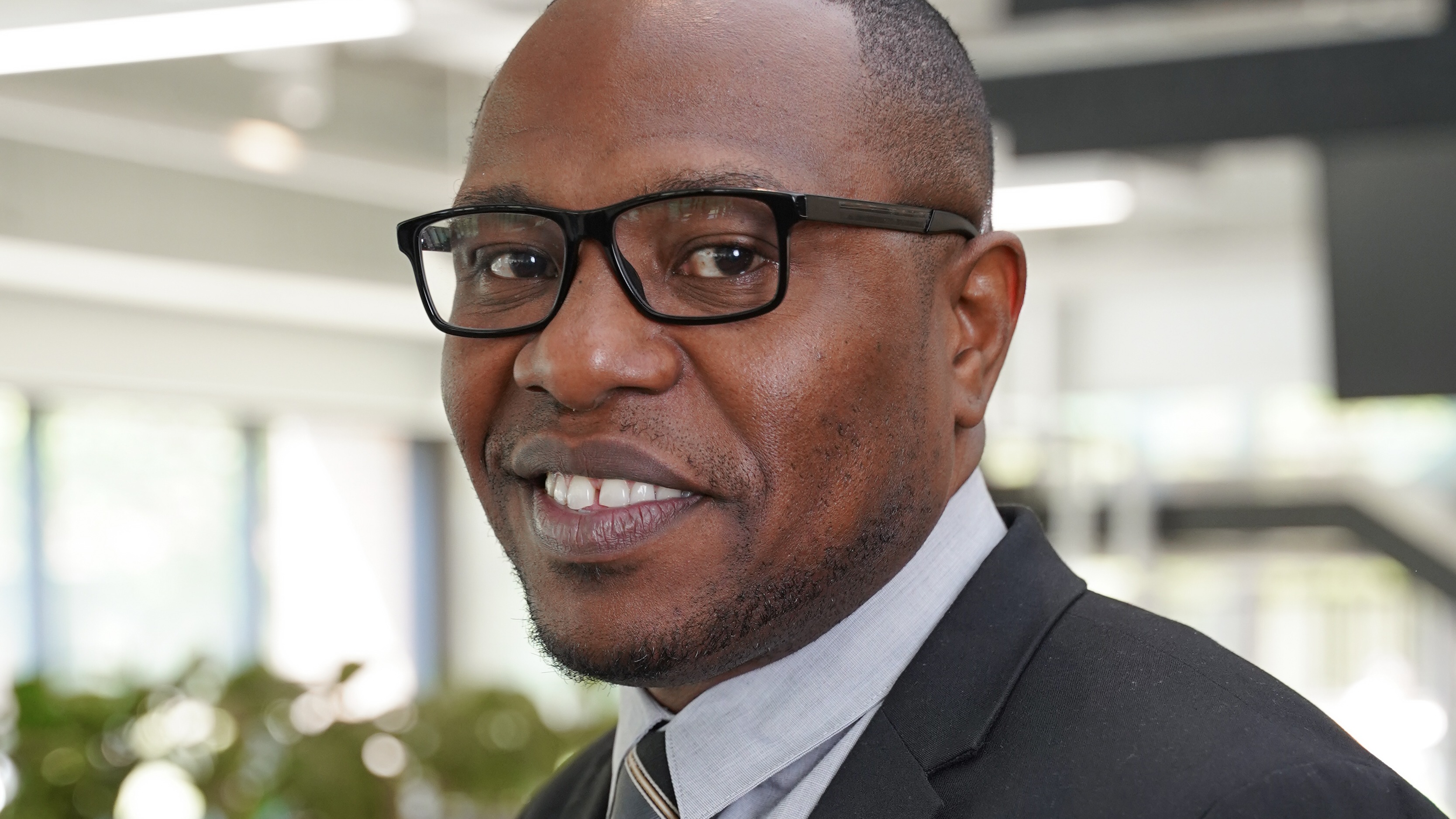 Karey Rowe wears glasses, a blazer, and tie as he looks into the camera smiling