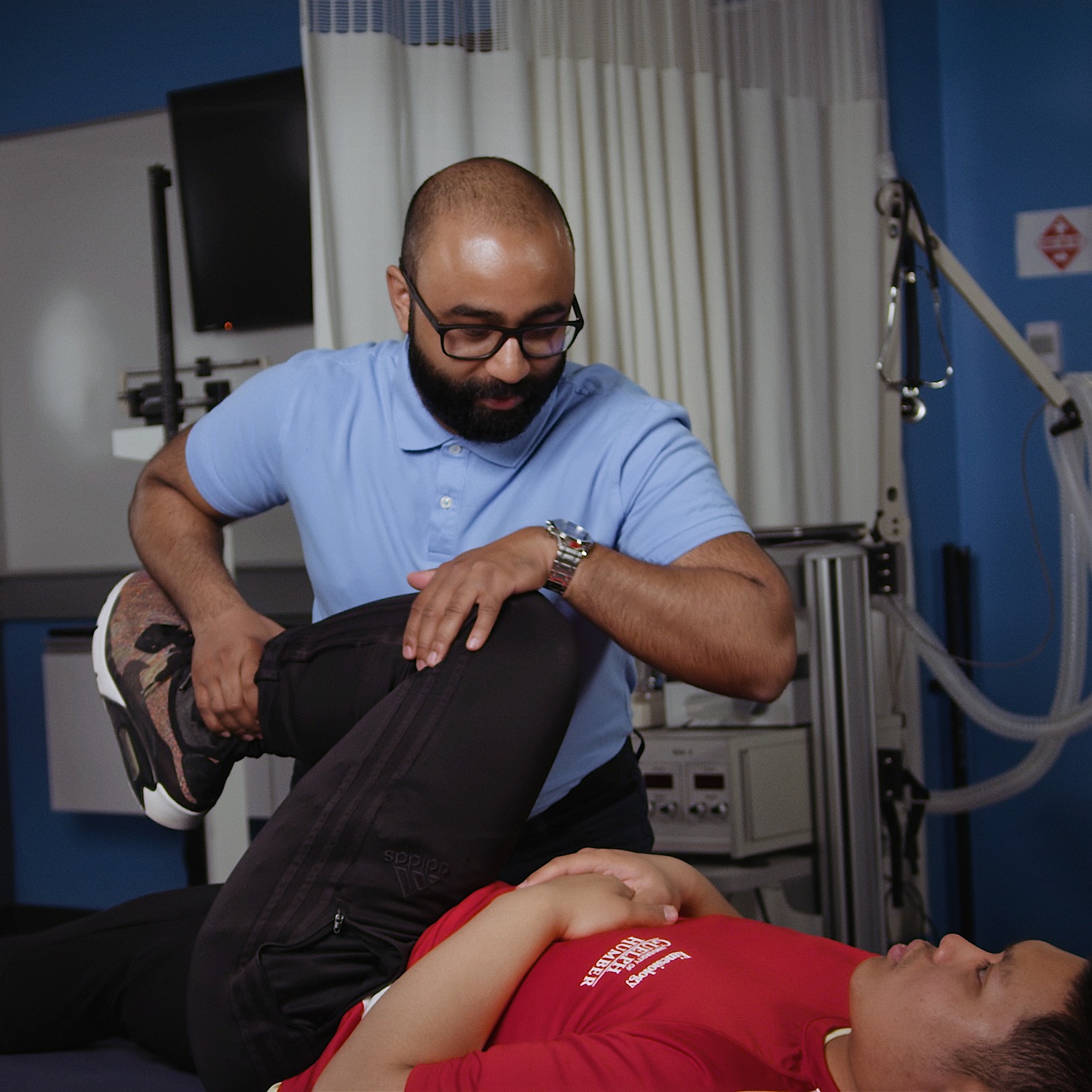 A man in a blue shirt holds the leg of a man in a red shirt, stretching him on a table