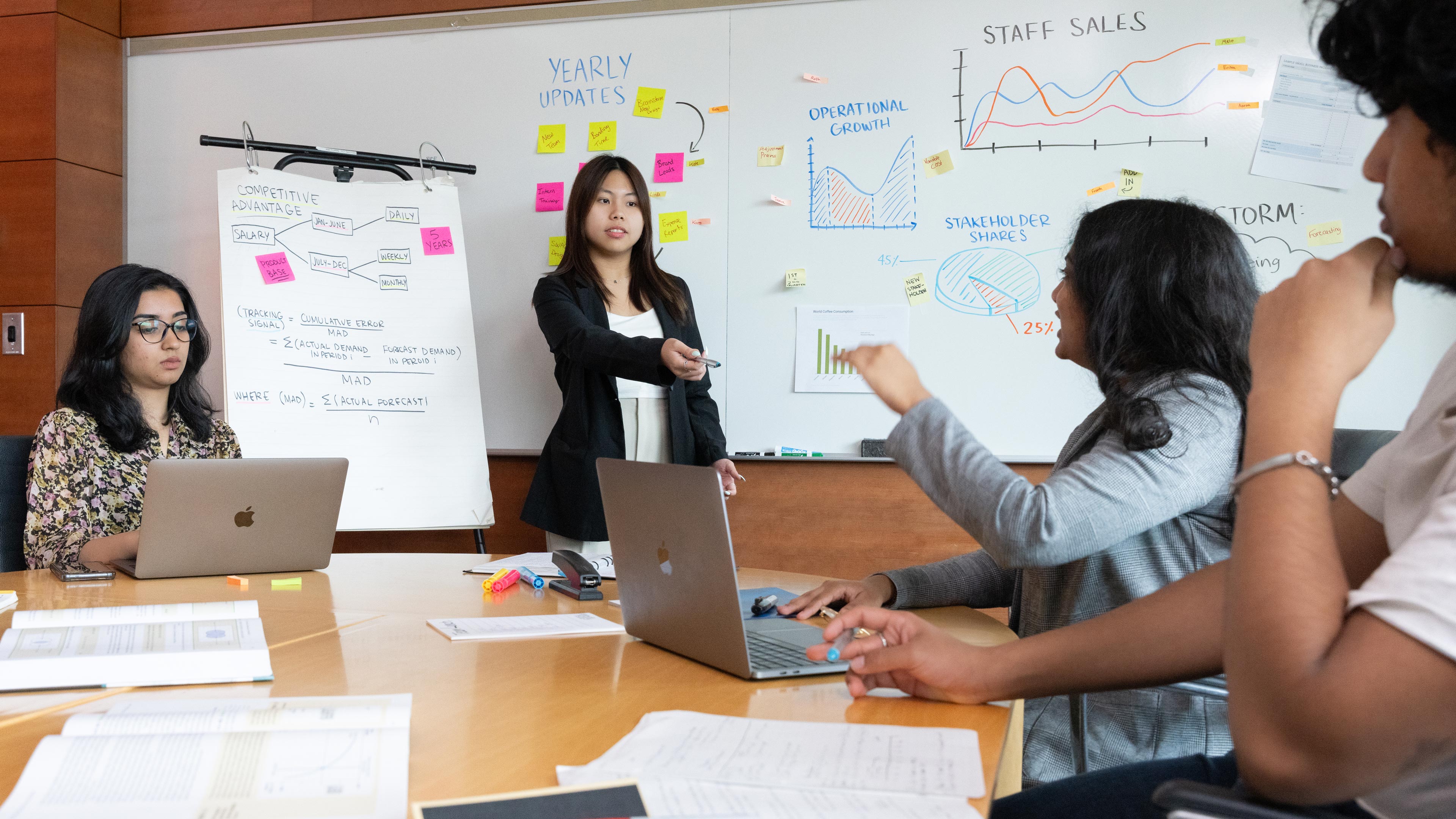 Students sit around a table with laptops, while another student stands in front of a white board pointing a pen