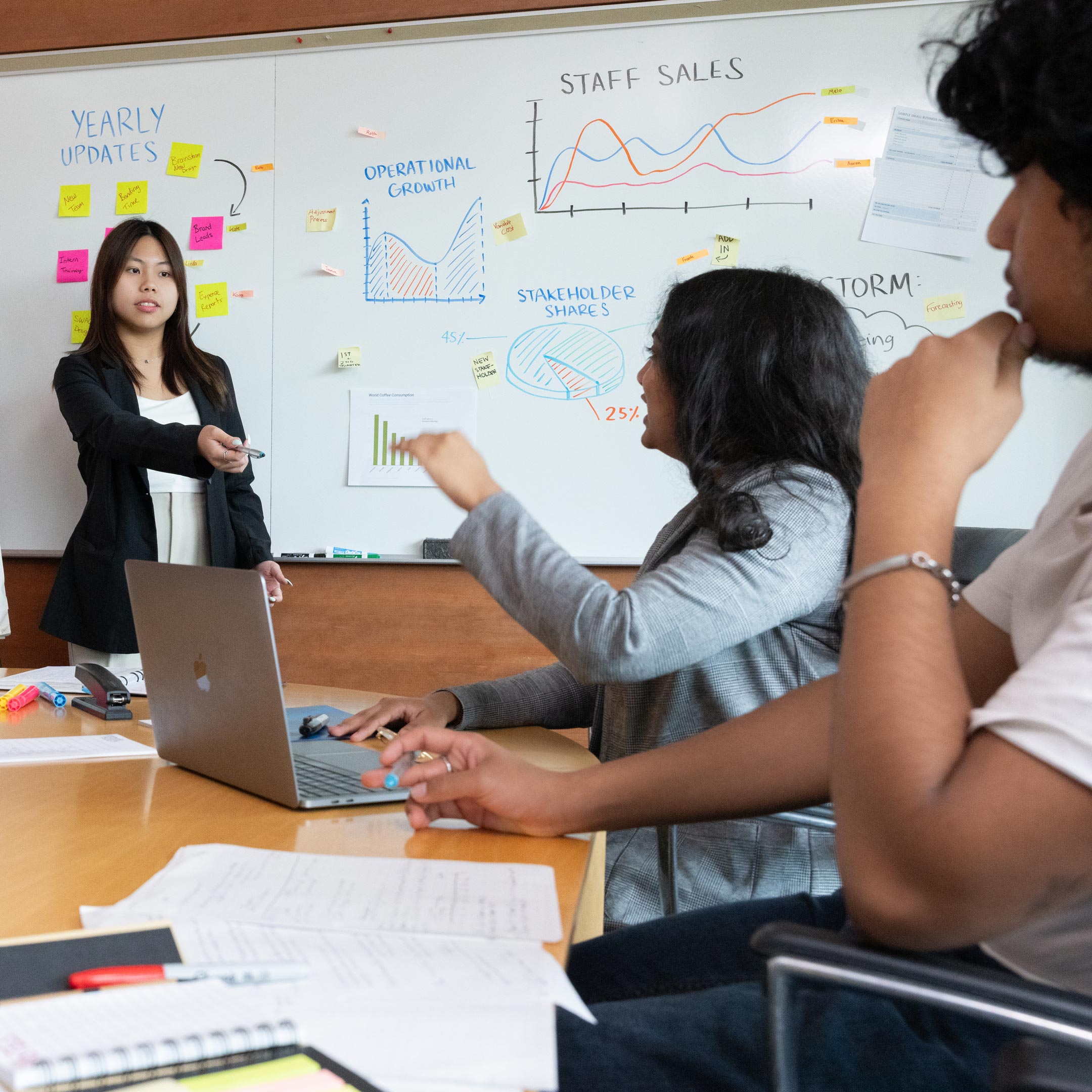 Students sit around a table with laptops, while another student stands in front of a white board pointing a pen