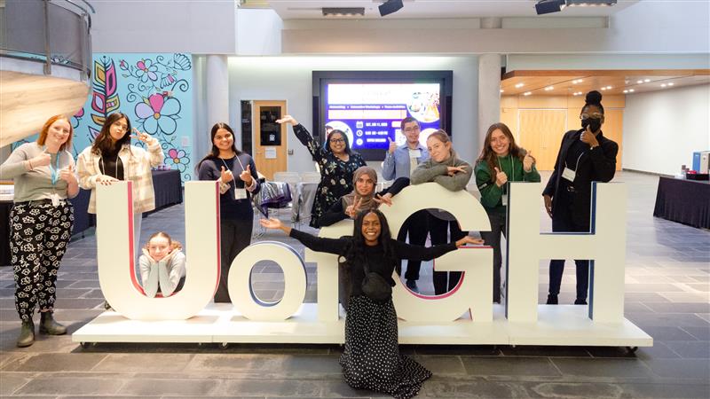 A group of students and staff pose in front of the large U of GH letters