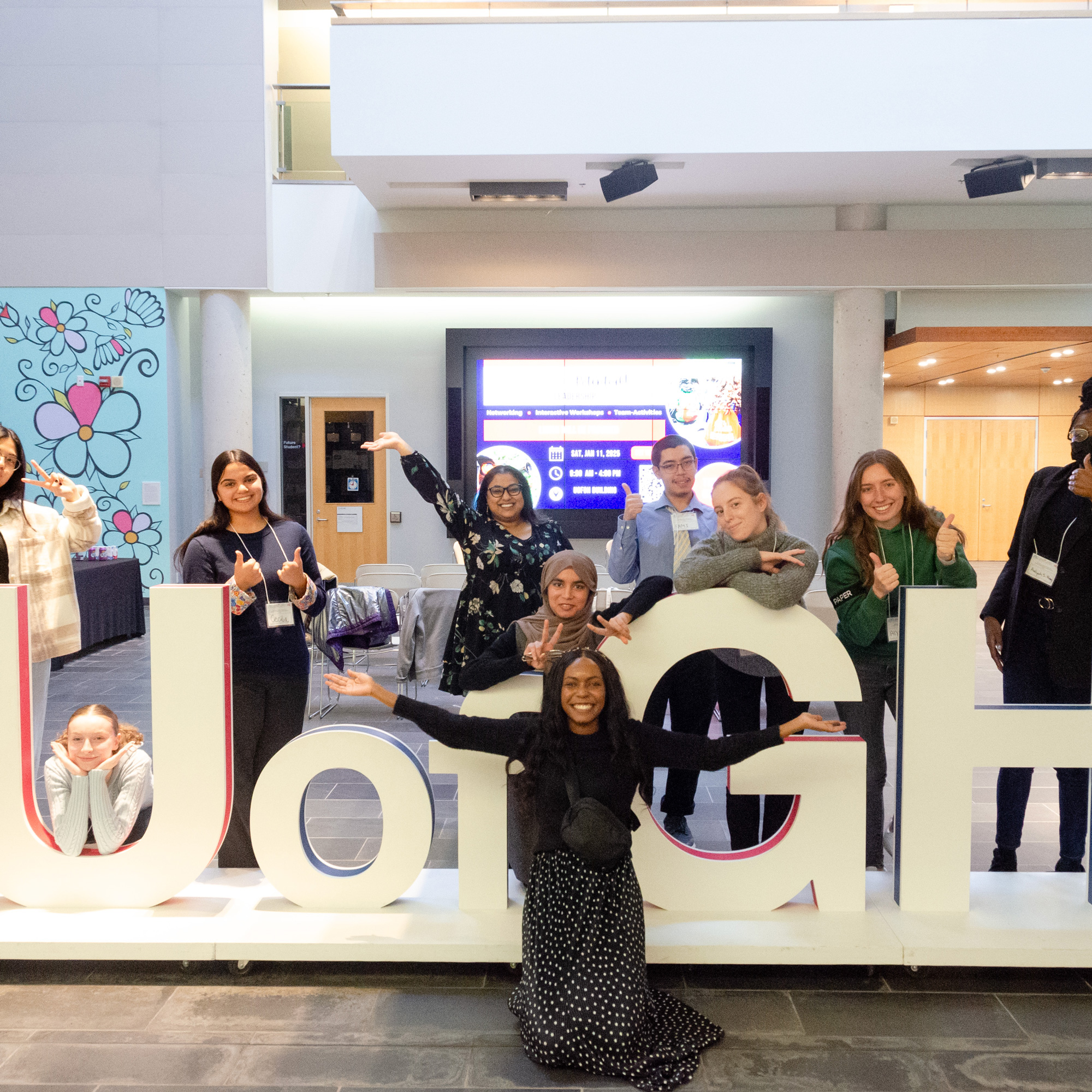 A group of students and staff pose in front of the large U of GH letters