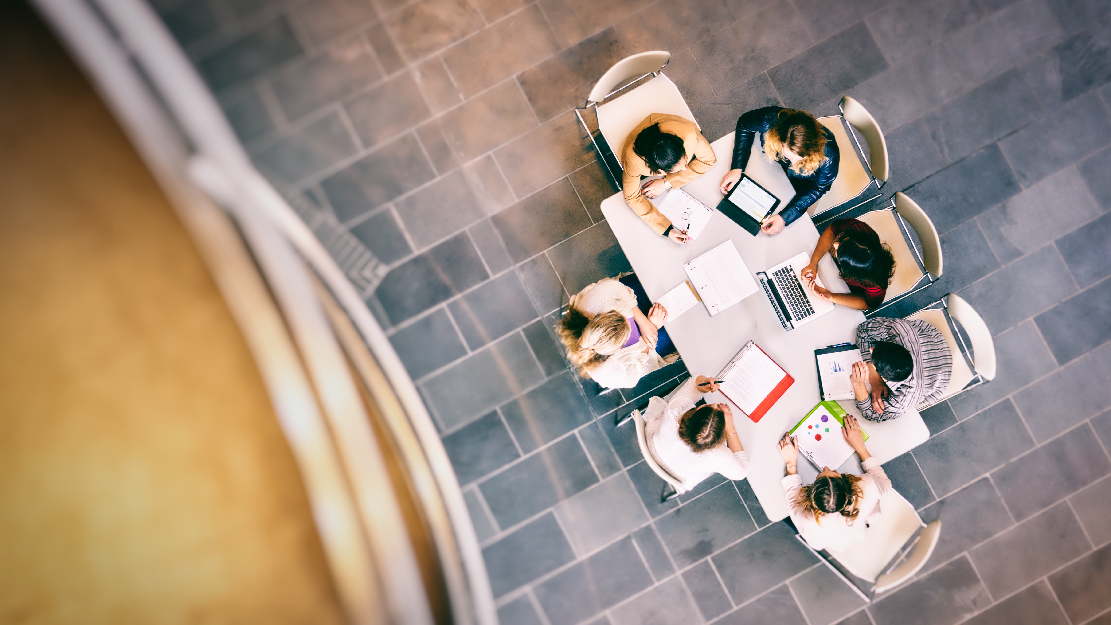 A group of seven students sit at a table gathering over books and laptops