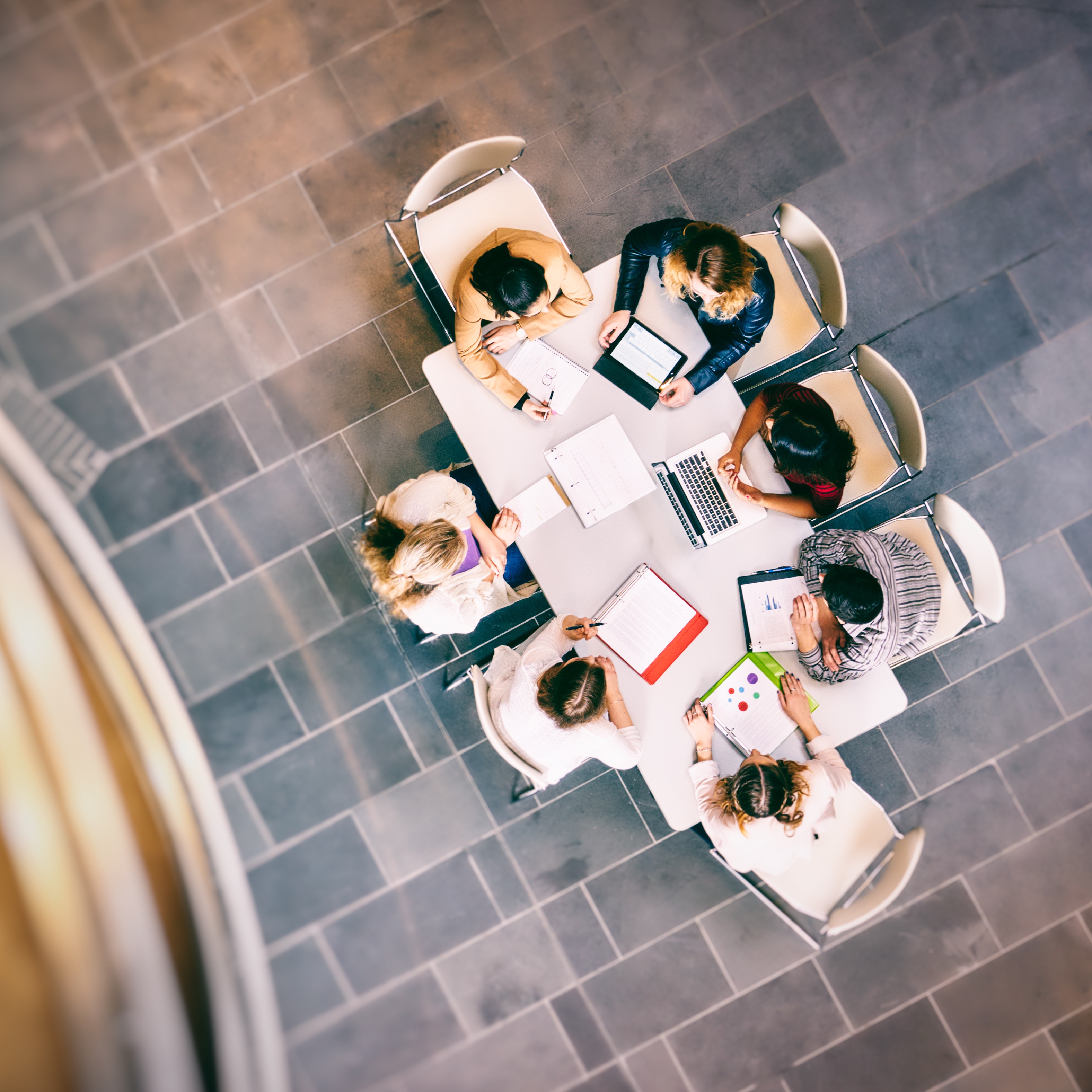 A group of seven students sit at a table gathering over books and laptops