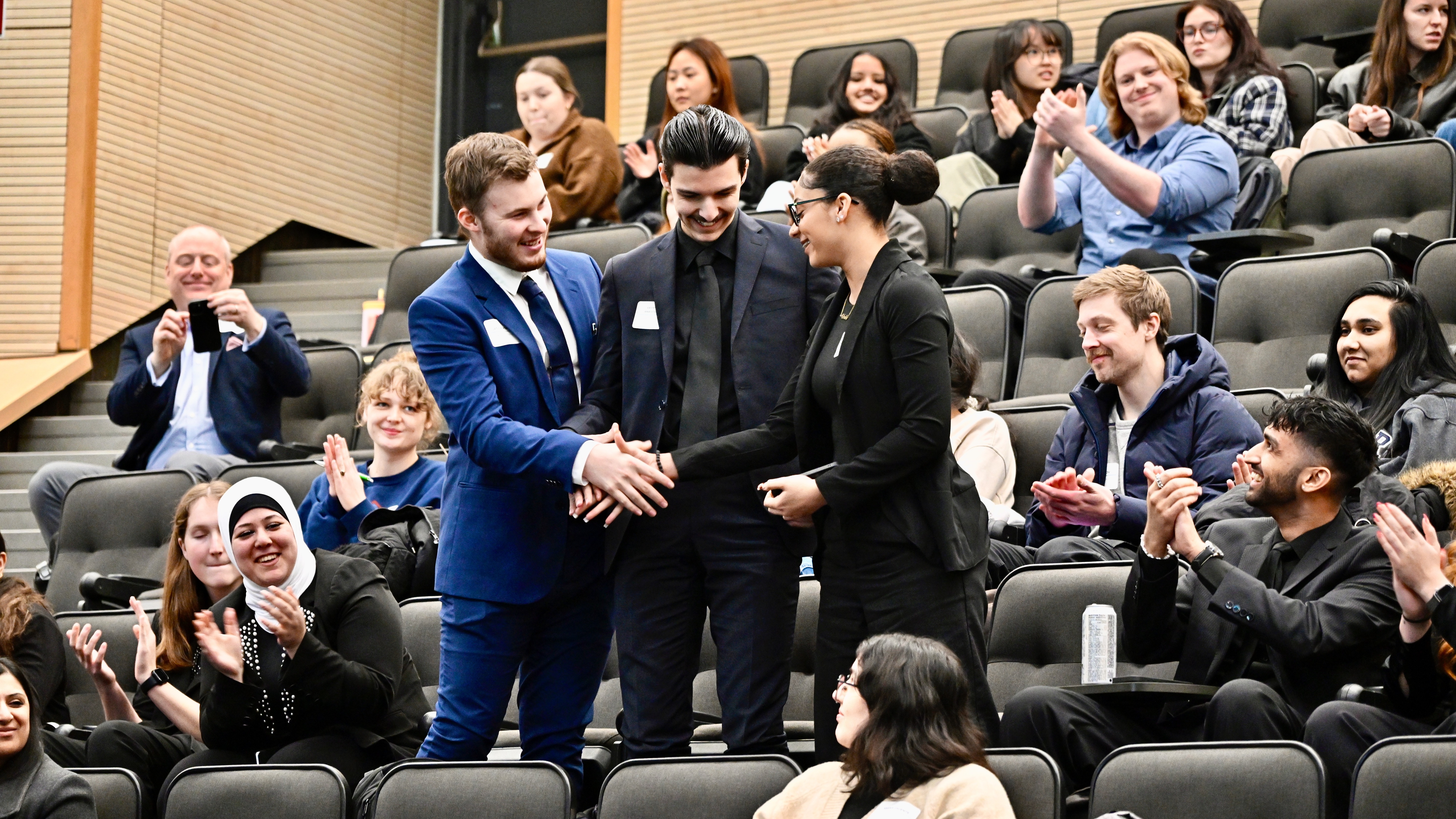 A group of three students shake hands among a crowd of applauding people