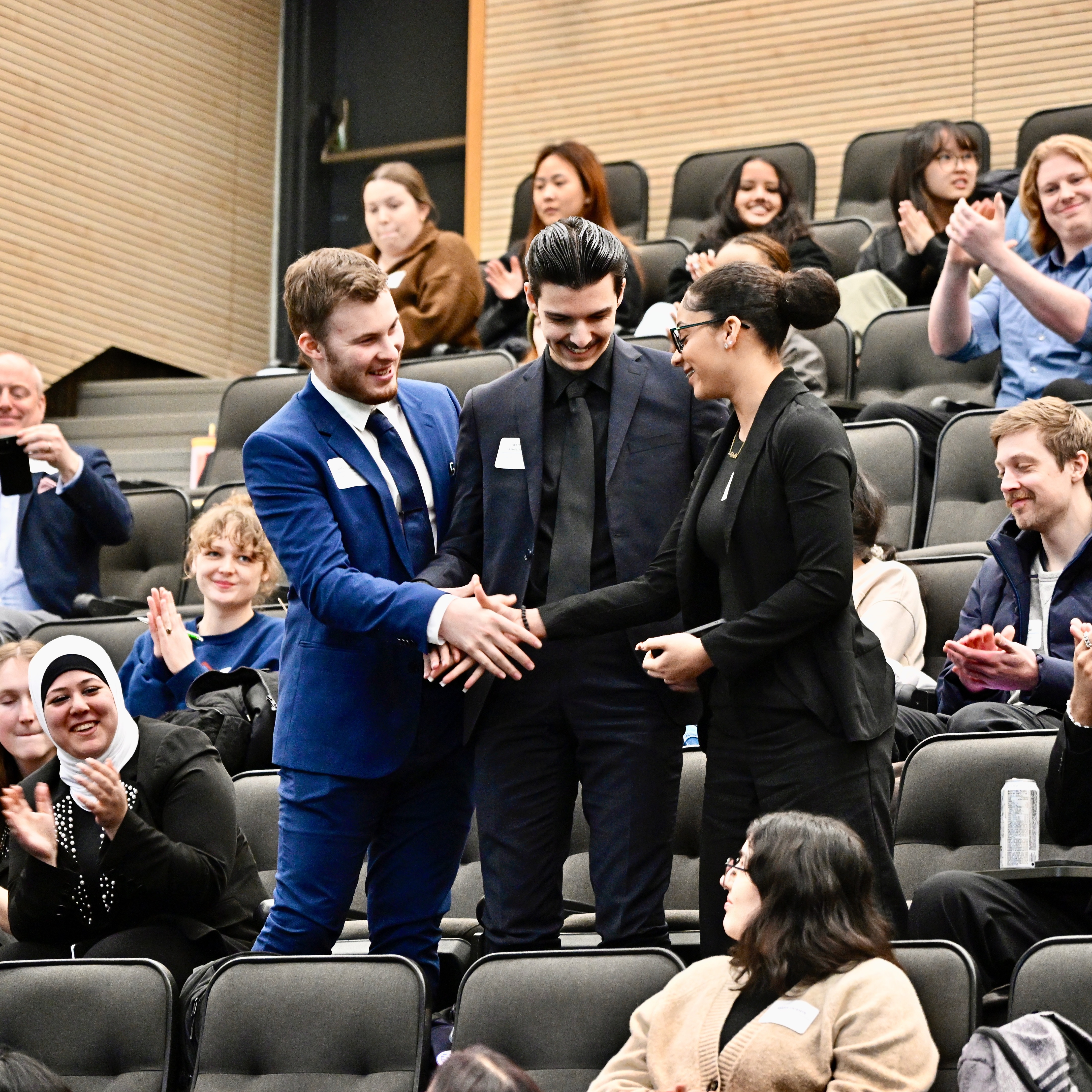 A group of three students shake hands among a crowd of applauding people