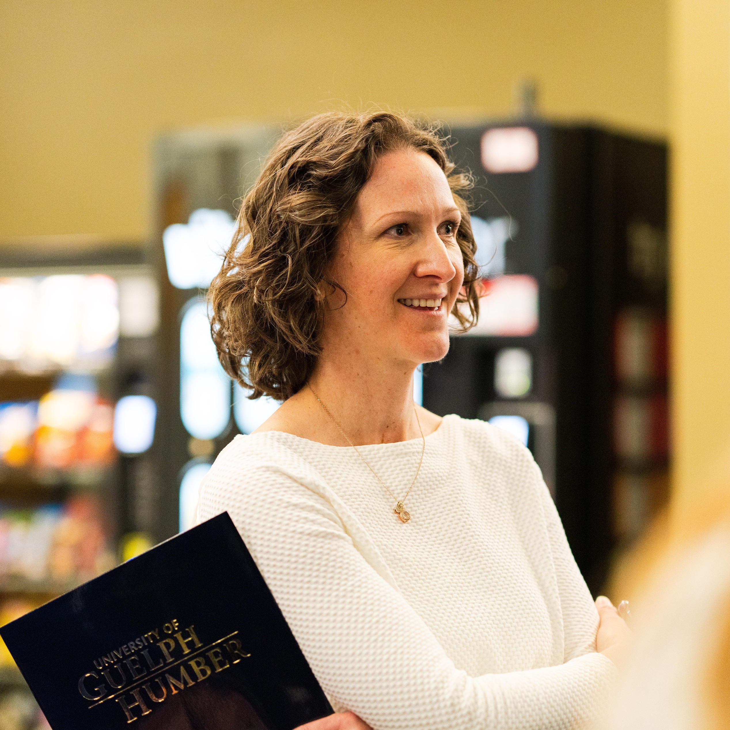 Dr. Leslie Auger wears a white top while holding a University of Guelph-Humber folder, looking to the side