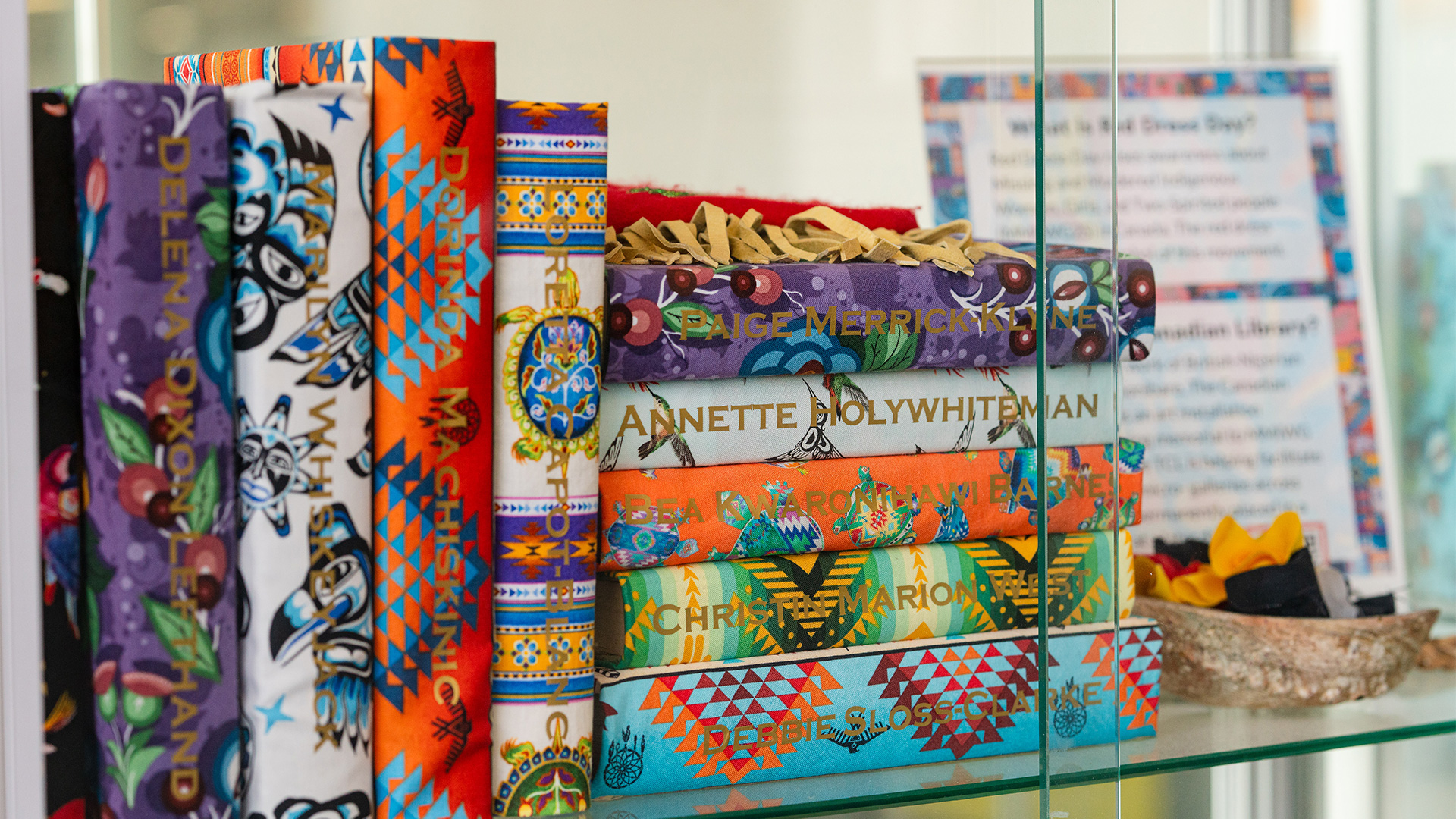 Books wrapped in colourful Indigenous fabrics with names of Missing and Murdered Indigenous Women, Girls, Two Spirit, and Gender-Diverse people and children on the spine are displayed on a glass shelf