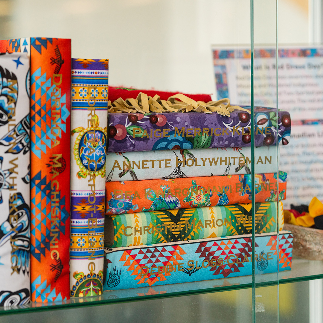 Books wrapped in colourful Indigenous fabrics with names of Missing and Murdered Indigenous Women, Girls, Two Spirit, and Gender-Diverse people and children on the spine are displayed on a glass shelf