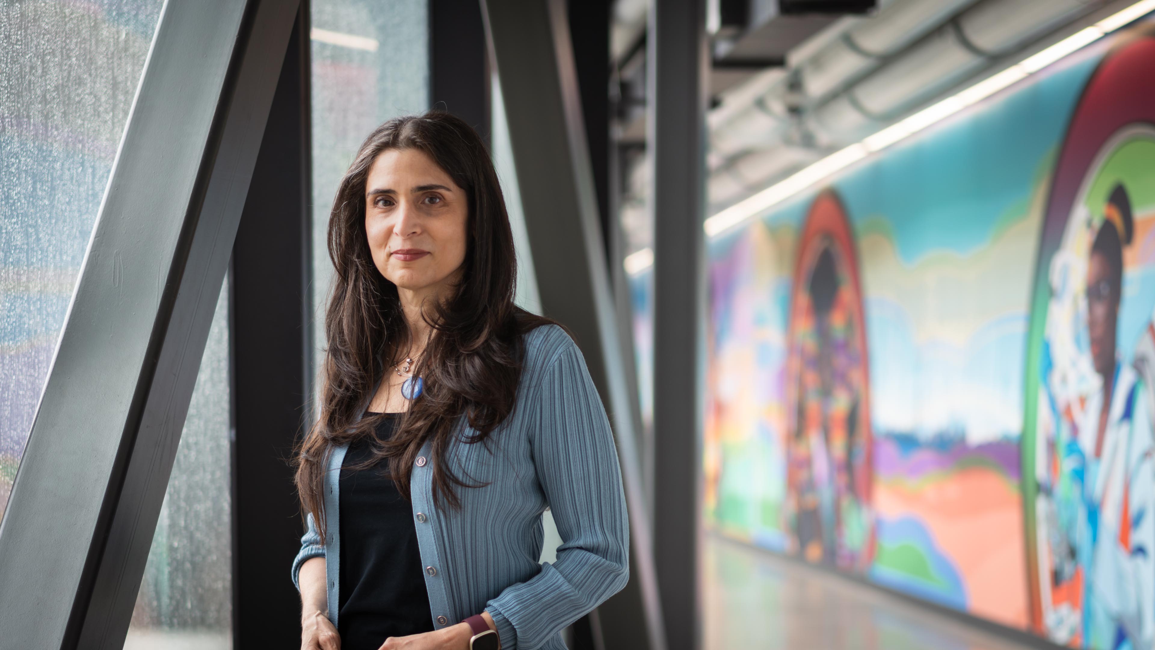 Dr. Khattar stands in a hallway with a colourful wall by a window, wearing a blue cardigan