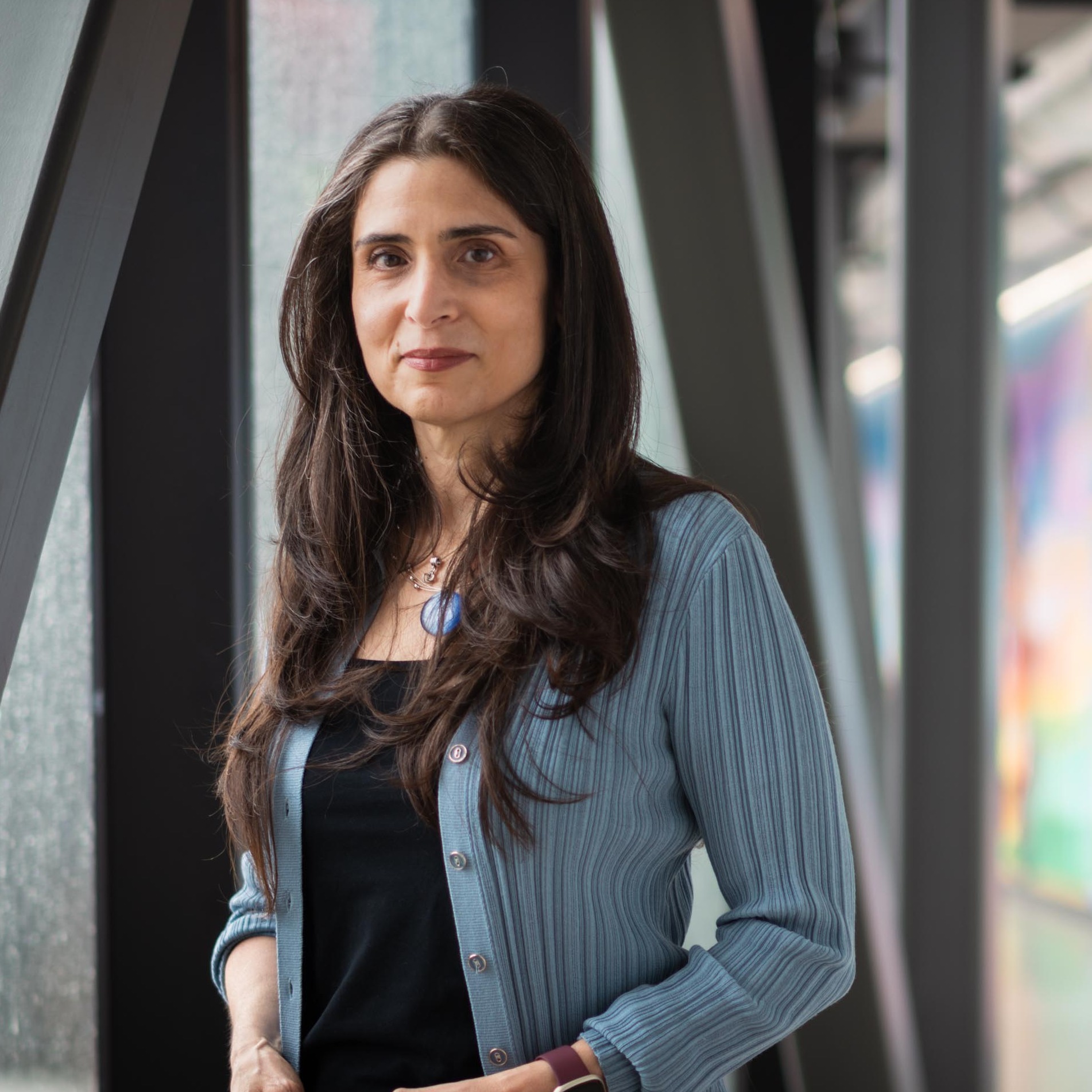 Dr. Khattar stands in a hallway with a colourful wall by a window, wearing a blue cardigan