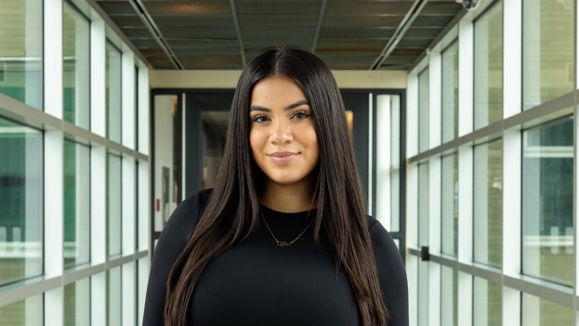 Alexia Velez wears a black top and stands in a window-filled hallway