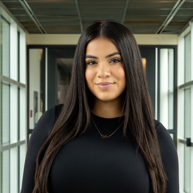 Alexia Velez wears a black top and stands in a window-filled hallway