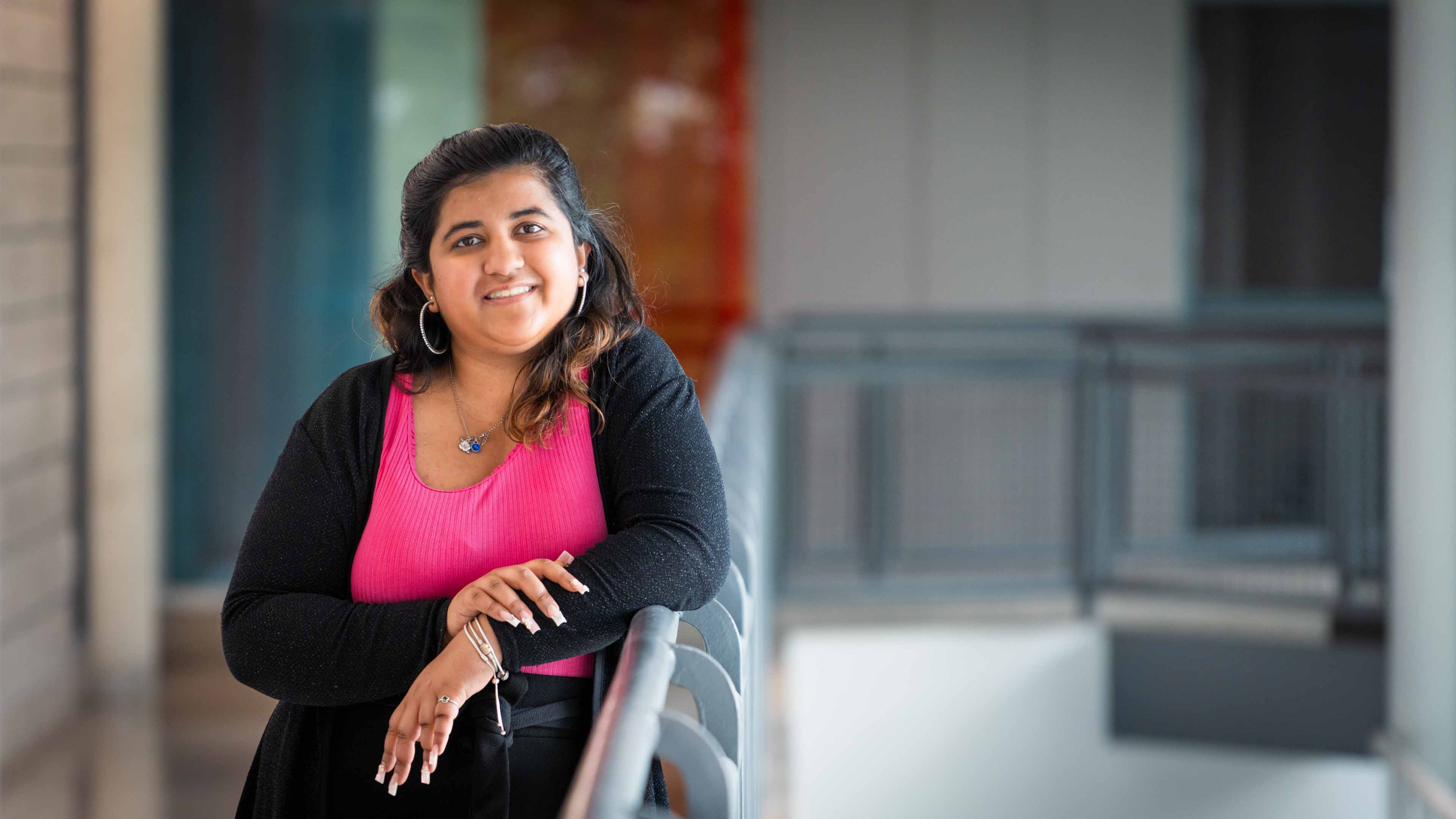 Nandita leans against a hand rail, wearing a bright pink top and black sweater