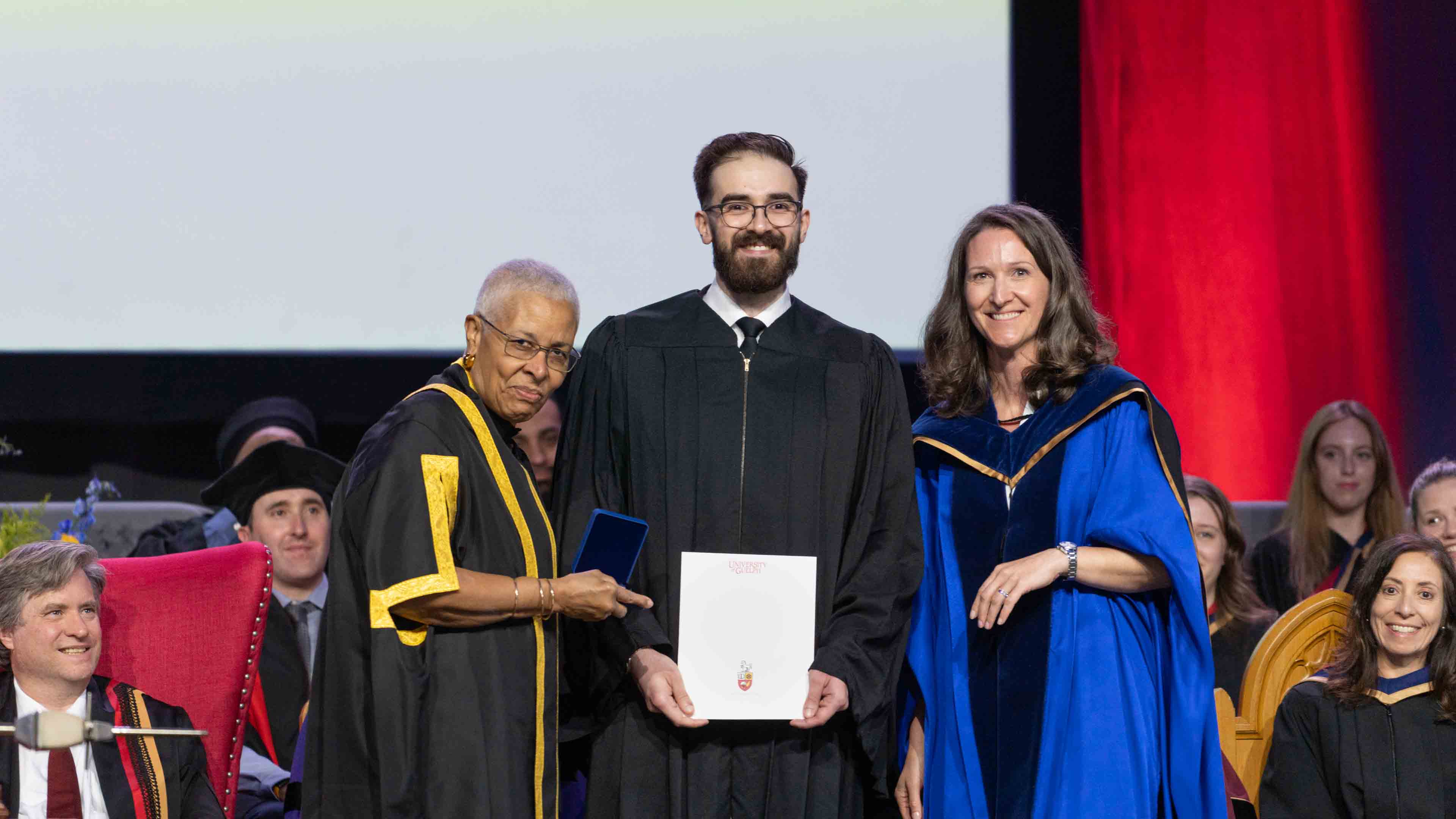Anthony Moncada poses on stage at convocation in his gown holding the honour alongside Dr. Chambers and Dr. Auger