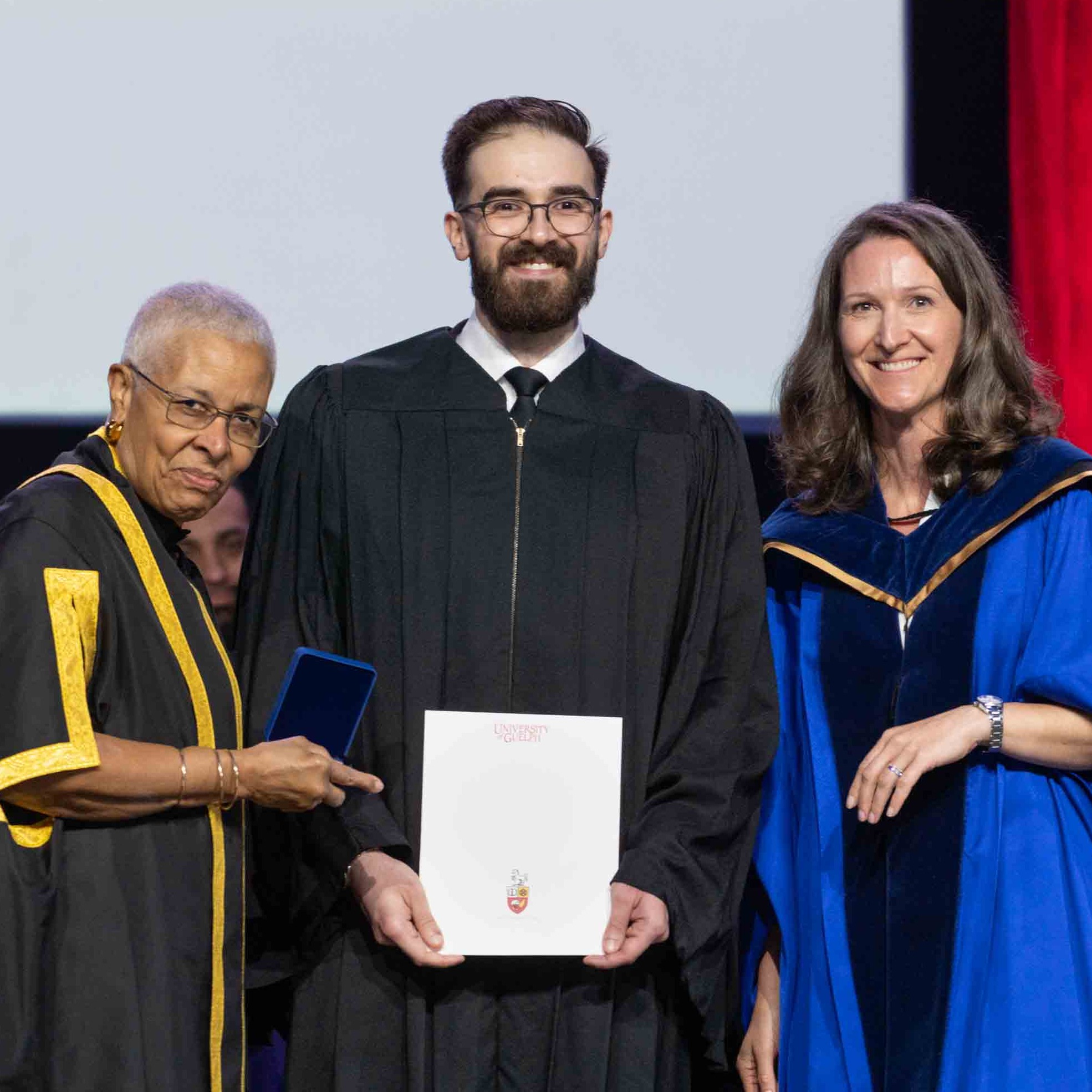 Anthony Moncada poses on stage at convocation in his gown holding the honour alongside Dr. Chambers and Dr. Auger