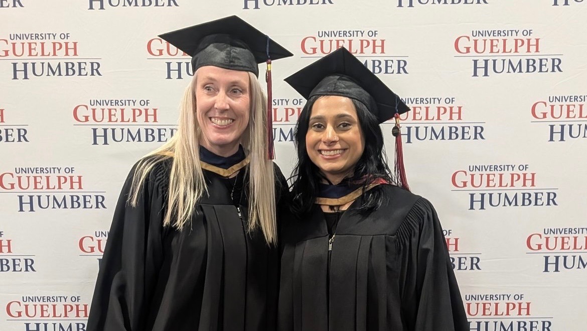 Two women wearing a graduate cap and gown pose in front of a University of Guelph-Humber banner