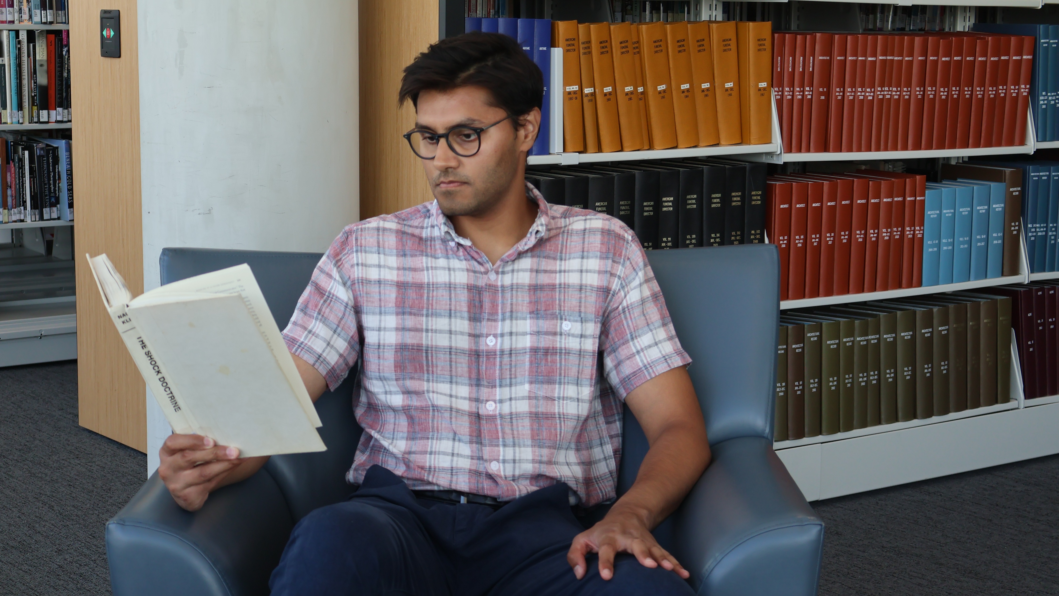 Usman Malik sits on a blue couch and reads a book in front of book shelves
