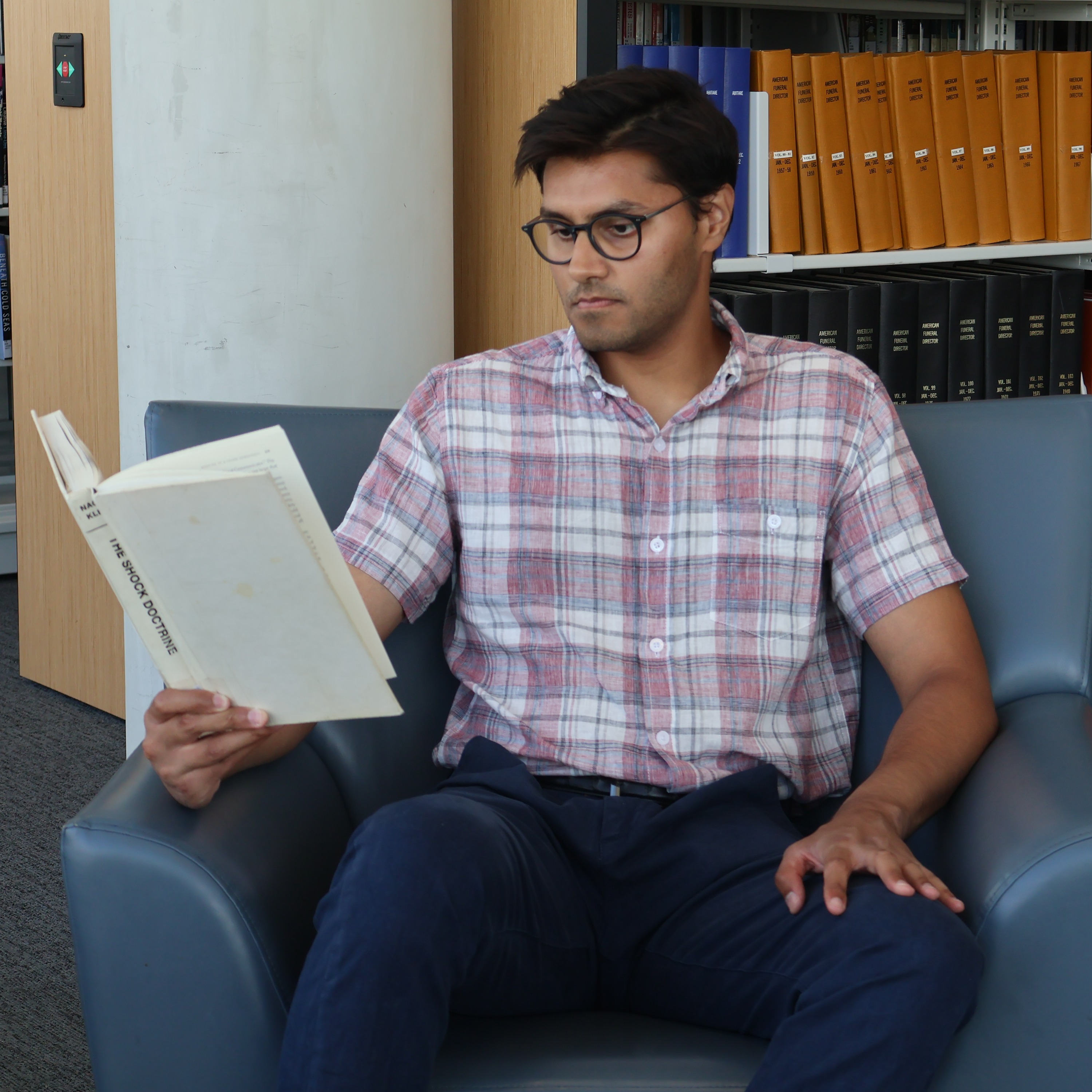 Usman Malik sits on a blue couch and reads a book in front of book shelves