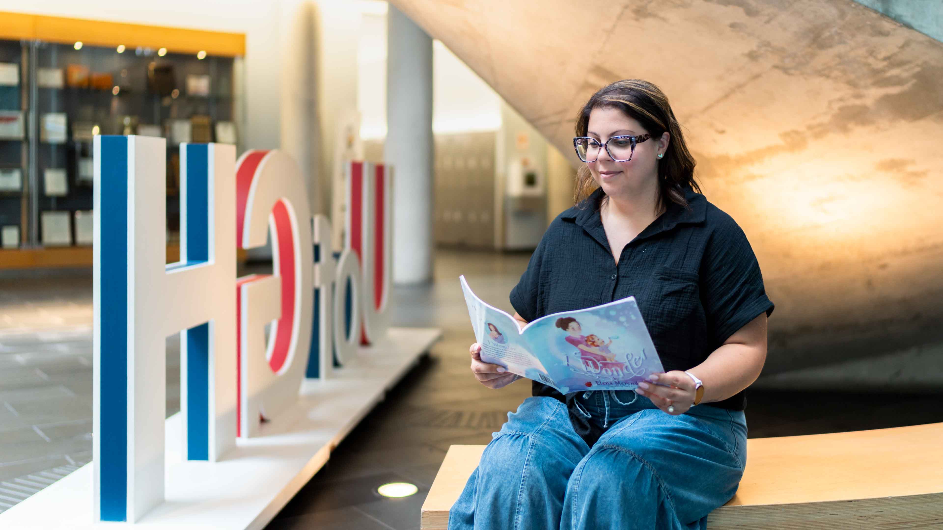 Dr. Elena Merenda sits on a bench, holding and reading her book, I Wonder, in the Guelph-Humber atrium