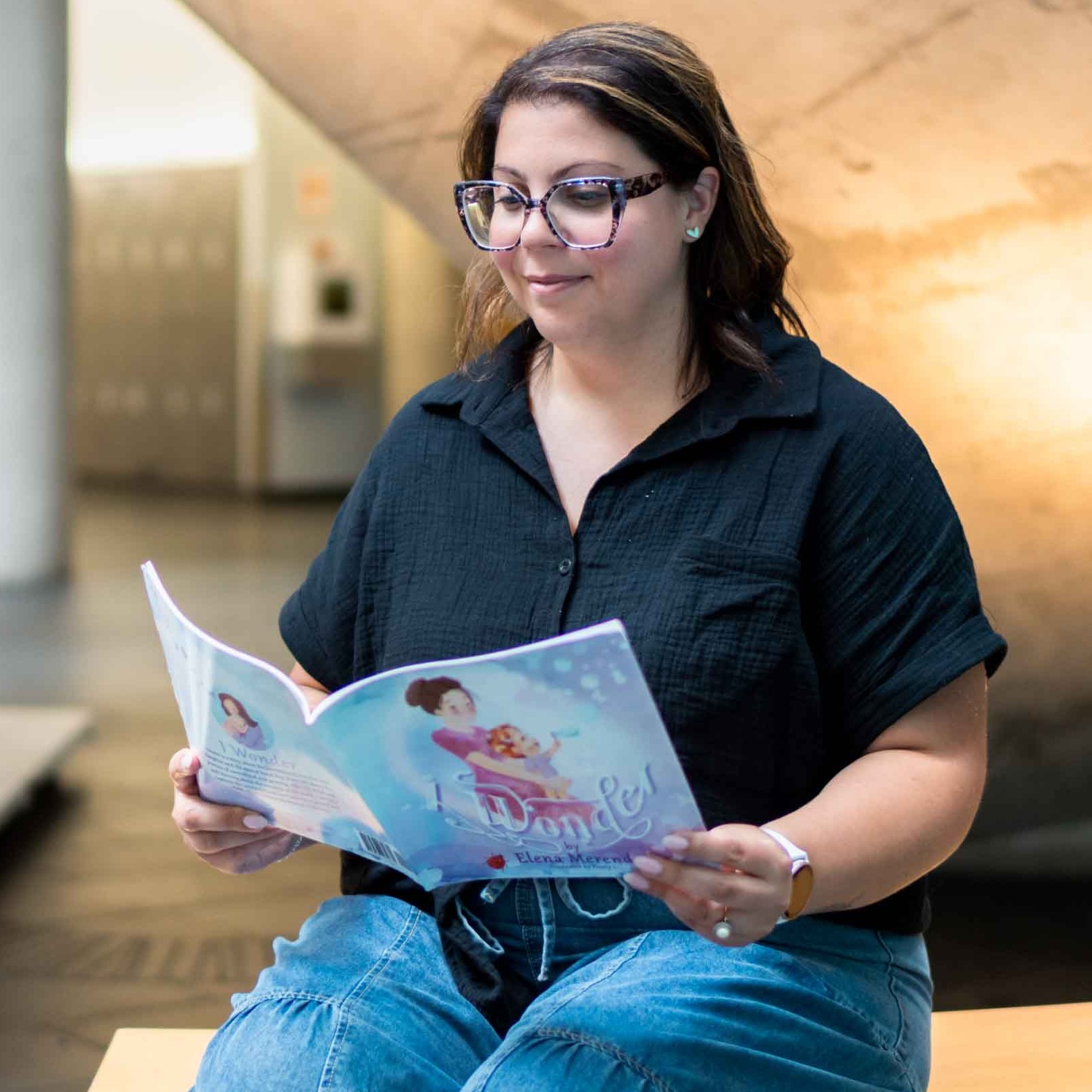 Dr. Elena Merenda sits on a bench, holding and reading her book, I Wonder, in the Guelph-Humber atrium
