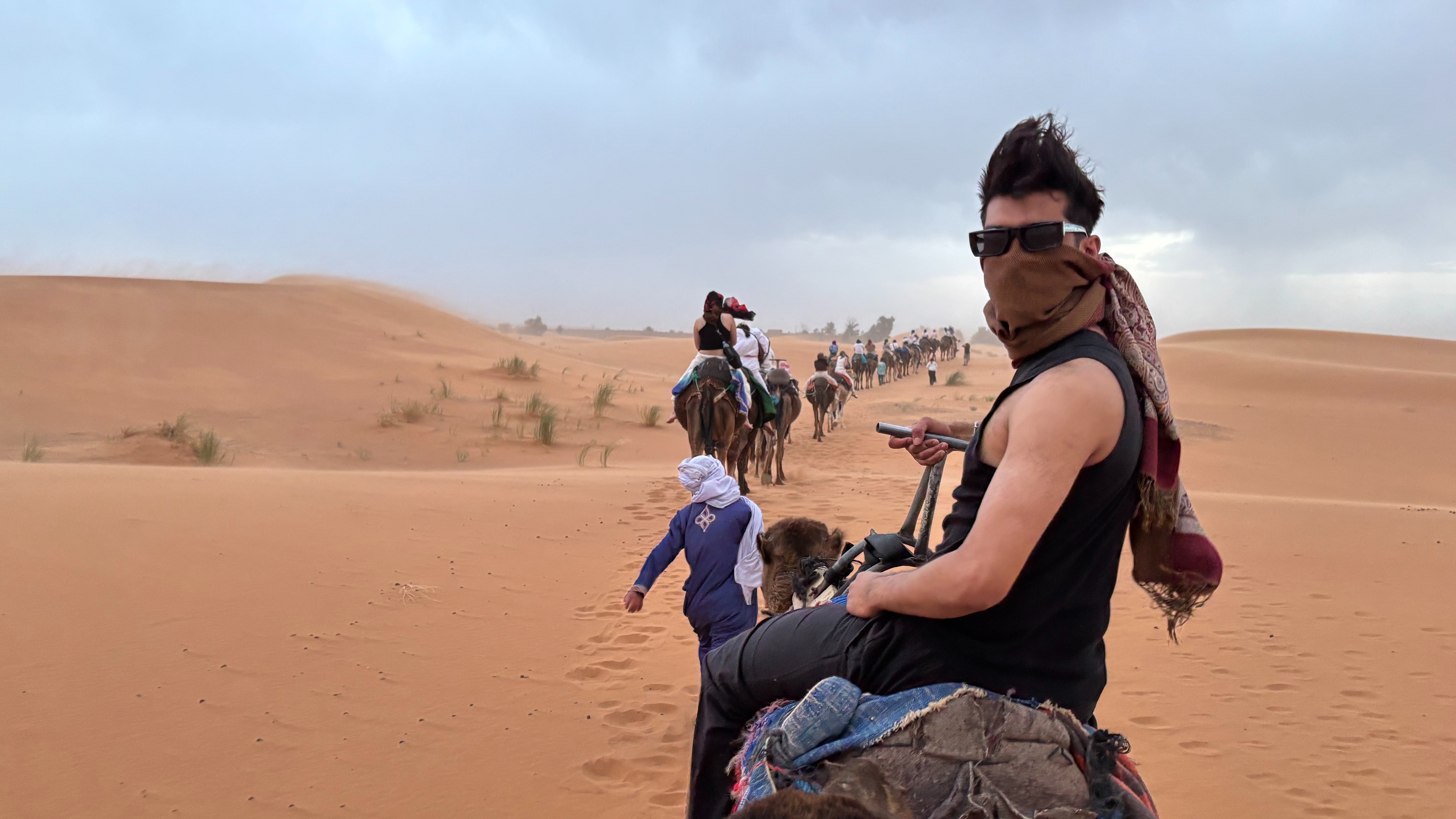 A student rides a camel in the Sahara Desert while wearing glasses and a scarf