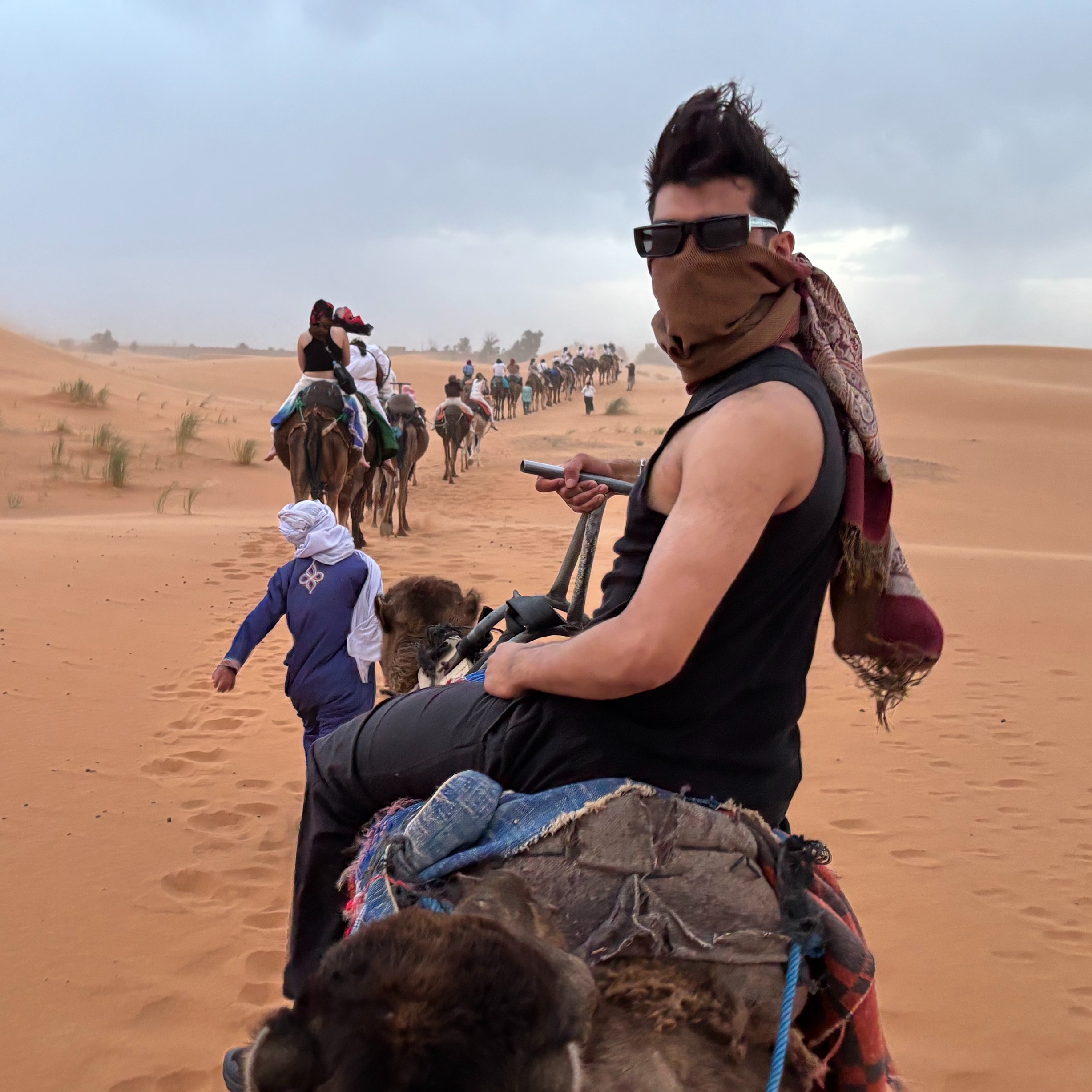 A student rides a camel in the Sahara Desert while wearing glasses and a scarf