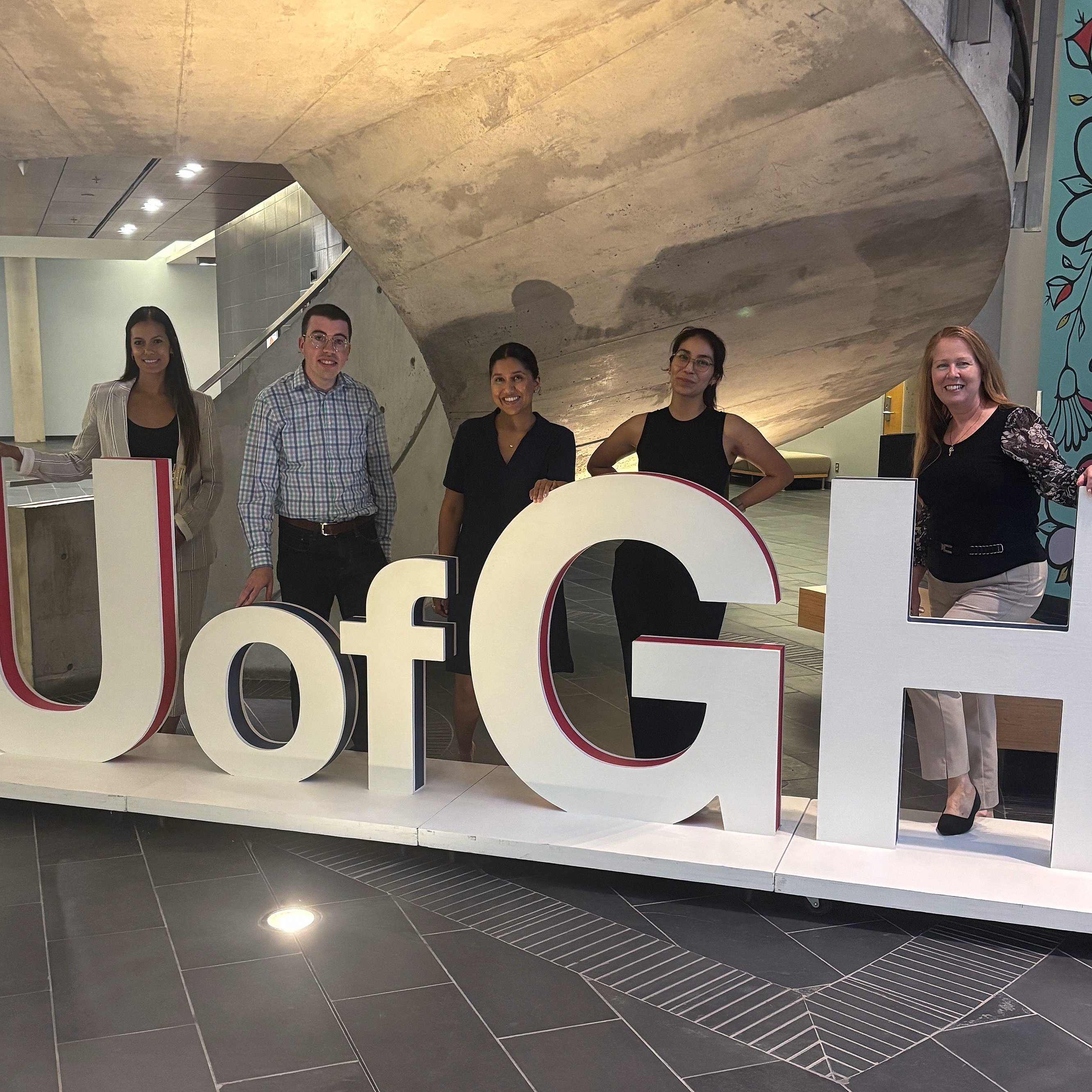 Alumni Ambassador Council members stand behind the giant U of GH letters under the spiral staircase in the atrium