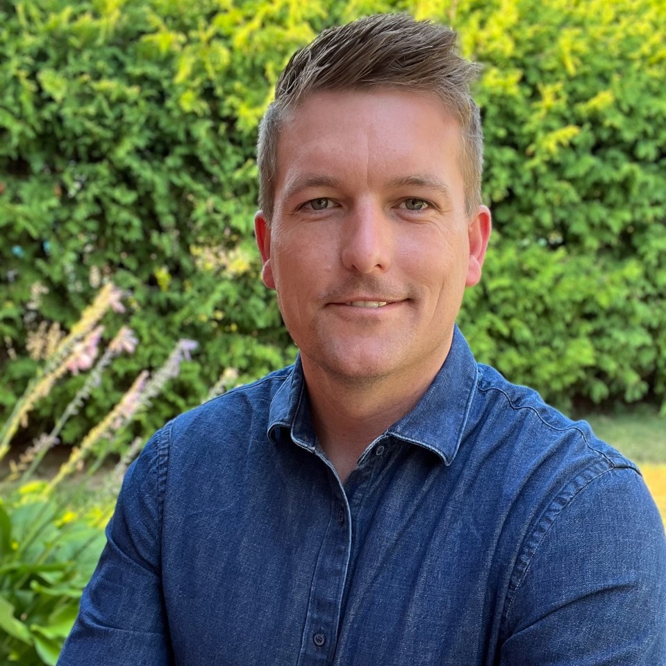 Alex Maveal smiles in front of a lush green garden