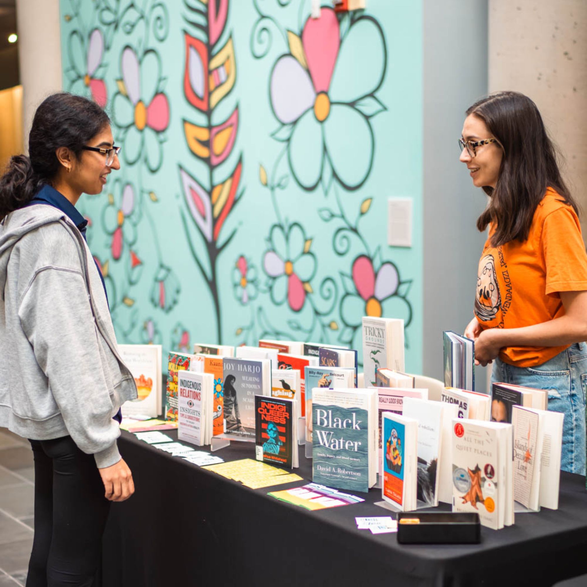 student at table with library books speaking with other student