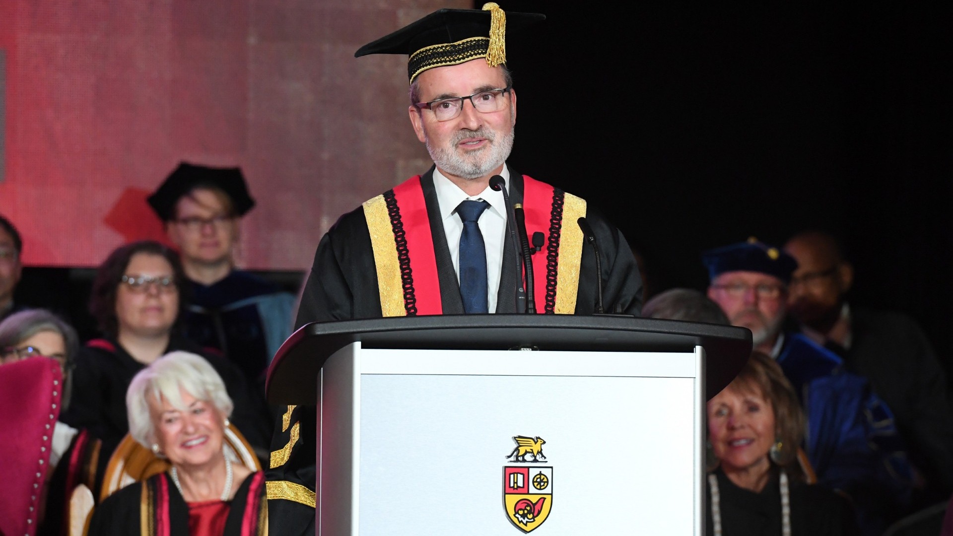 Dr. Van Acker speaks at a podium in academic regalia