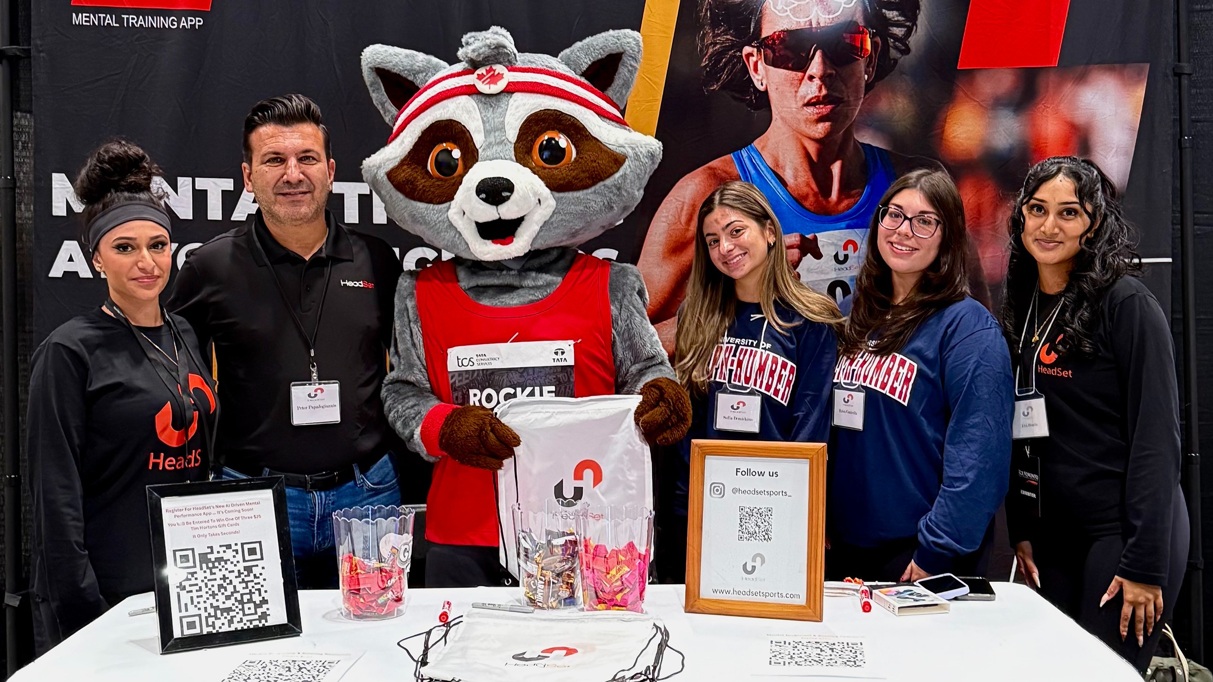 A group of students, professionals, and instructor Peter Papadogiannis pose with a mascot at their booth during the Toronto Waterfront Marathon Expo