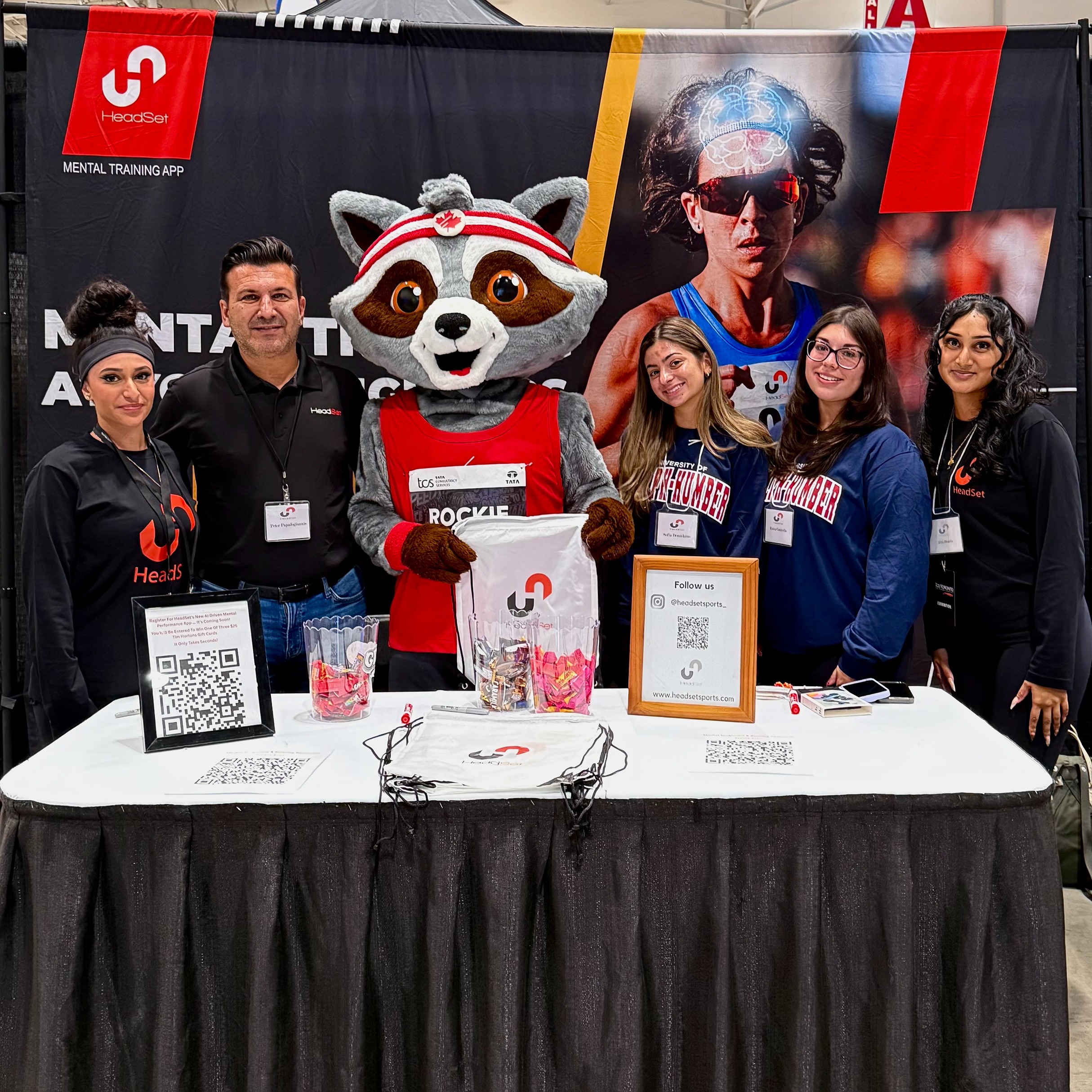 A group of students, professionals, and instructor Peter Papadogiannis pose with a mascot at their booth during the Toronto Waterfront Marathon Expo