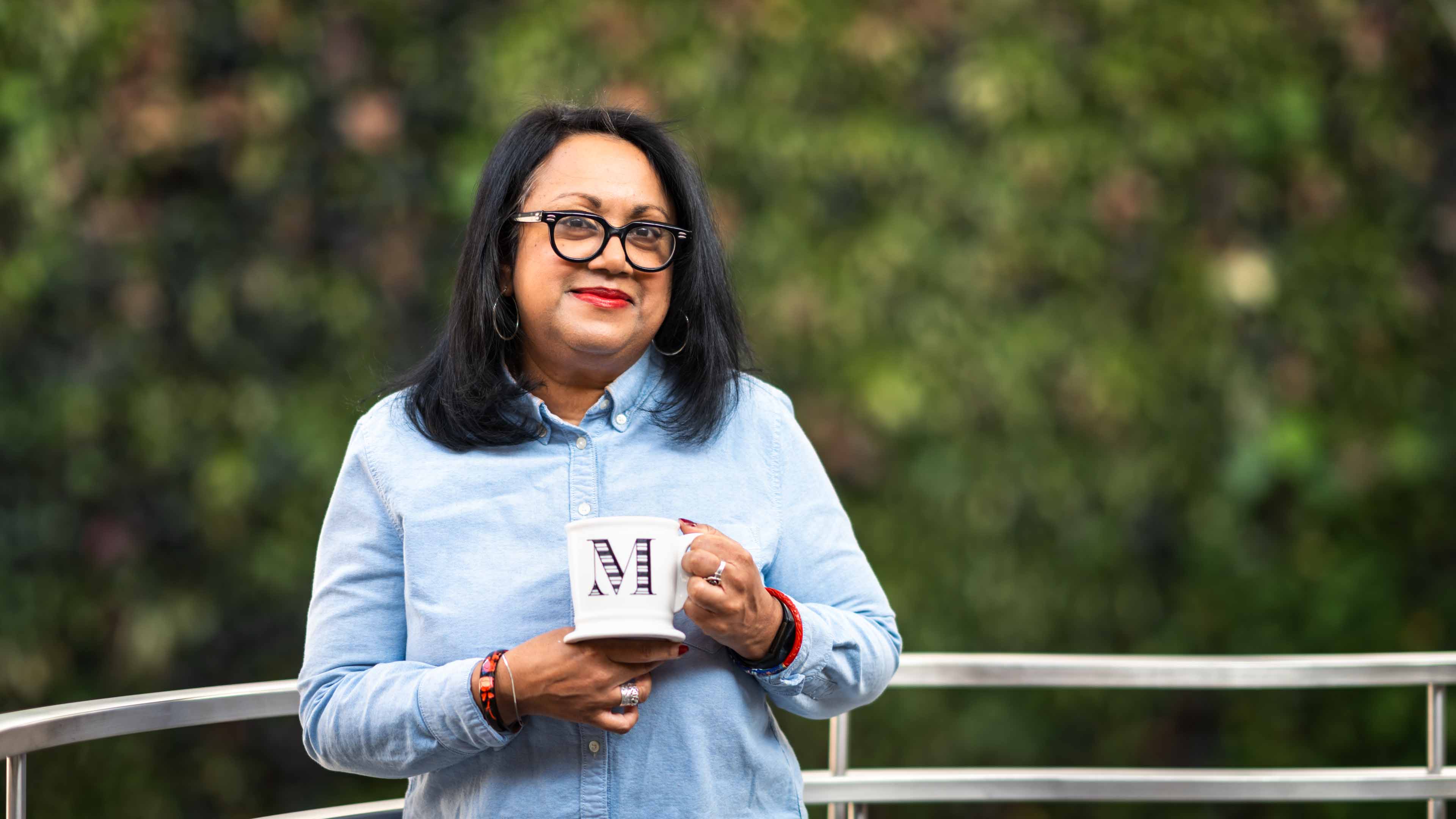 Melanie Zuzarte stands in front of a plant wall while holding a mug with the letter M on it