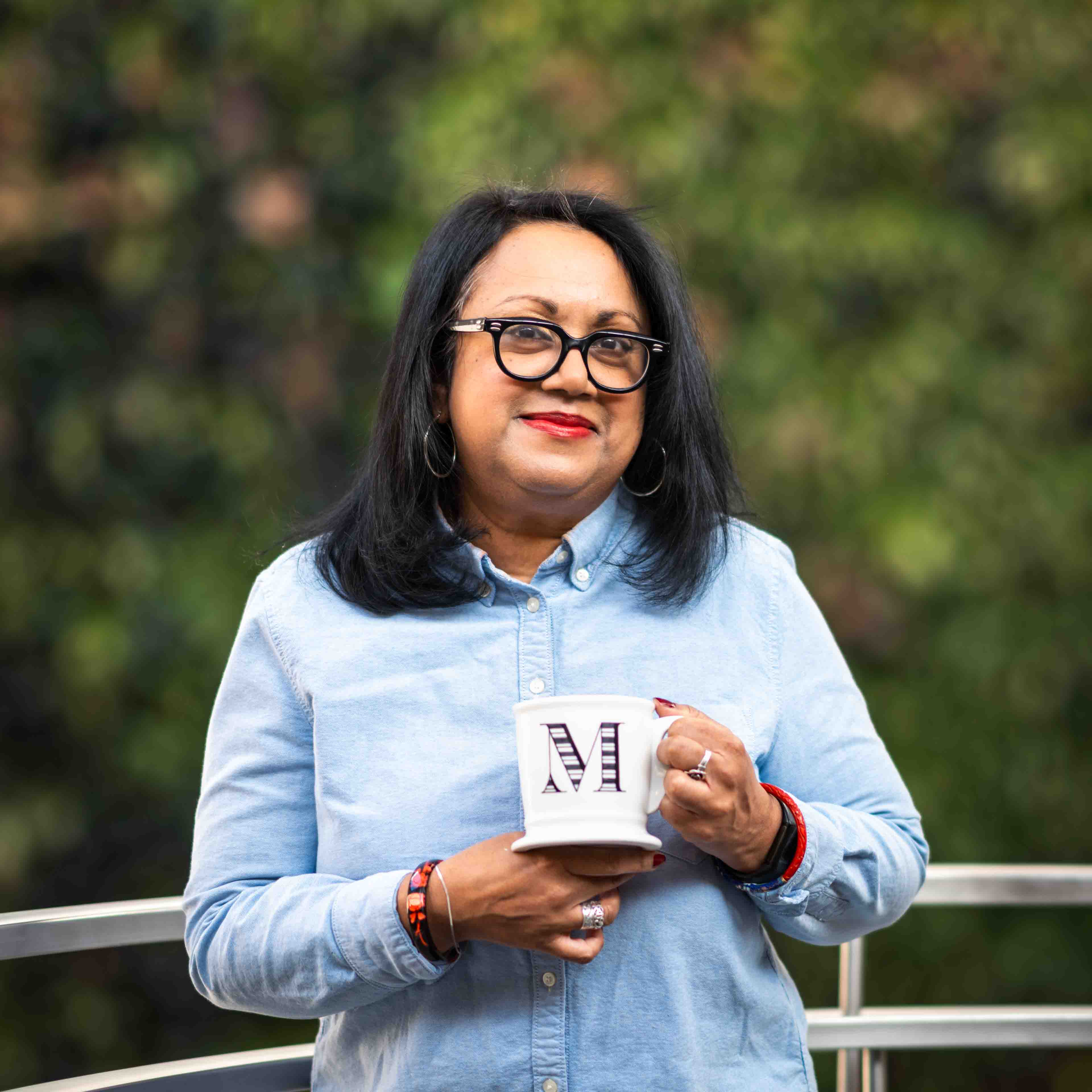 Melanie Zuzarte stands in front of a plant wall while holding a mug with the letter M on it